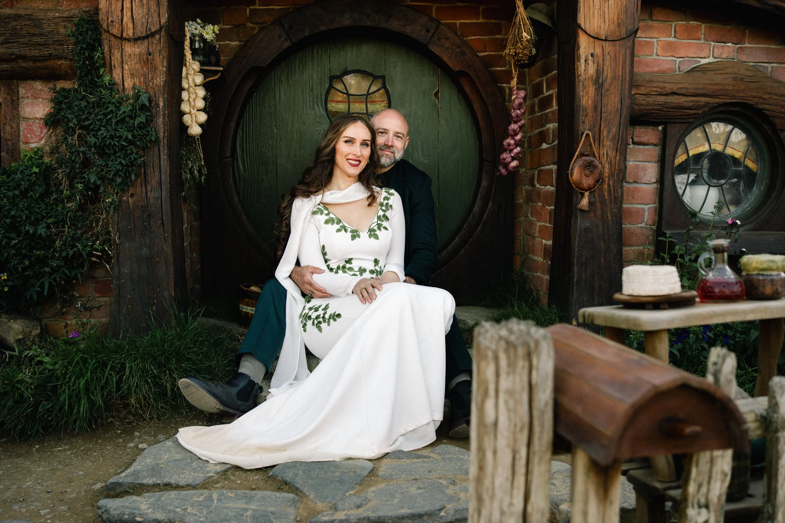 beautiful American couple siting infront of a round green door at Hobbiton movie set, they are smiling, she is sitting in-between his legs, there us foliage around them and a small wooden letterbox in the foreground out of focus