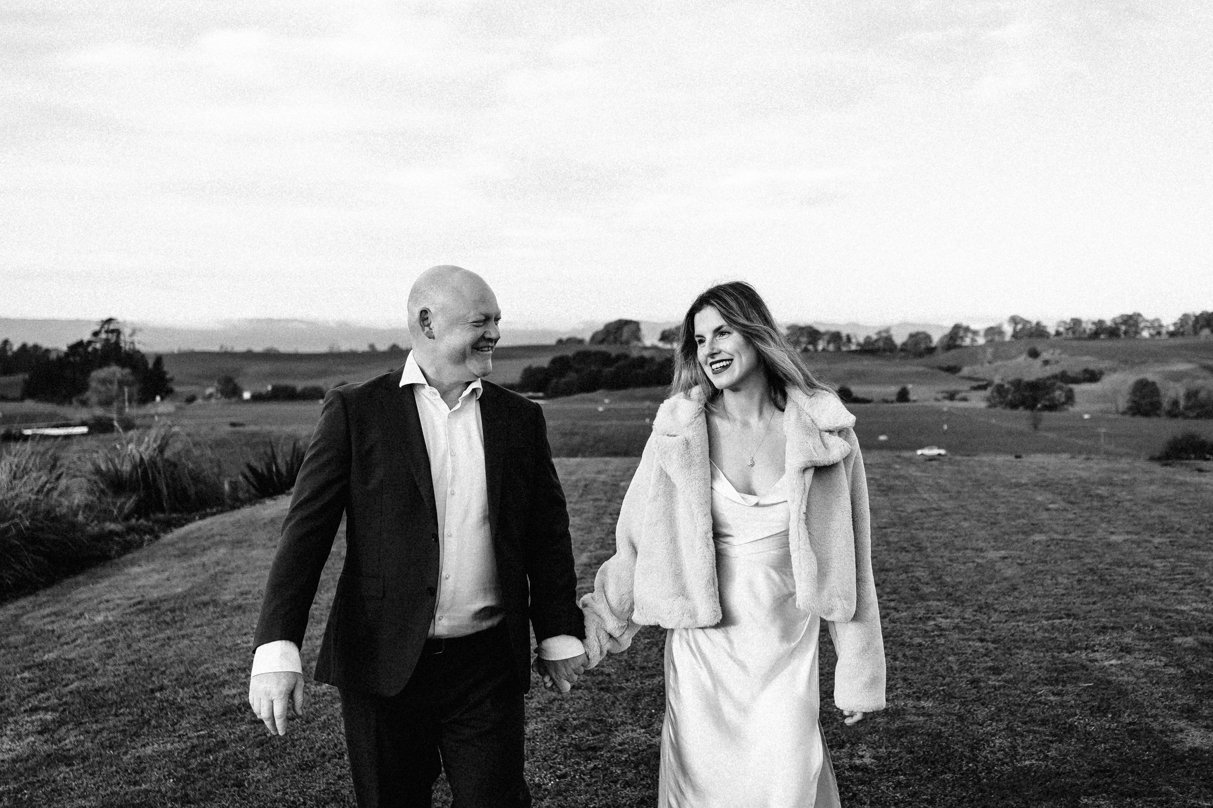 Bride in a long white silk wedding dress with a fur jacket is holding groom hand who in black suit, with white shirt, smiling, in a green field