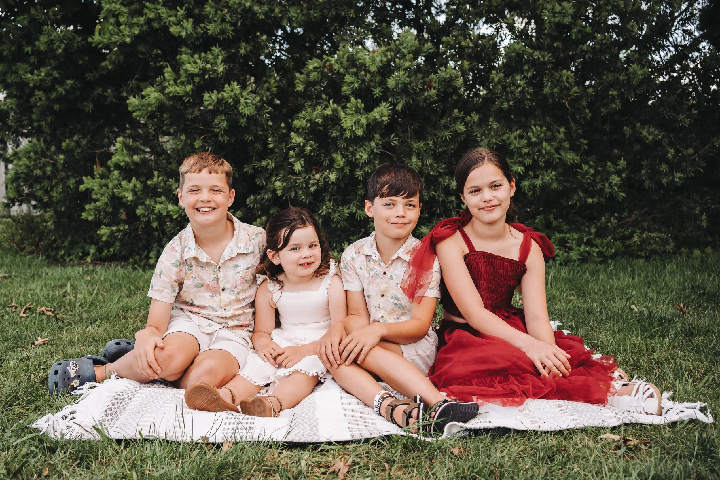 four siblings, two sisters and two brothers are sitting side by side on a cream rug on the grass with bushy foliage in the background, they area all smiling and looking at the camera, they are wearing a mix of cream tones and muted red