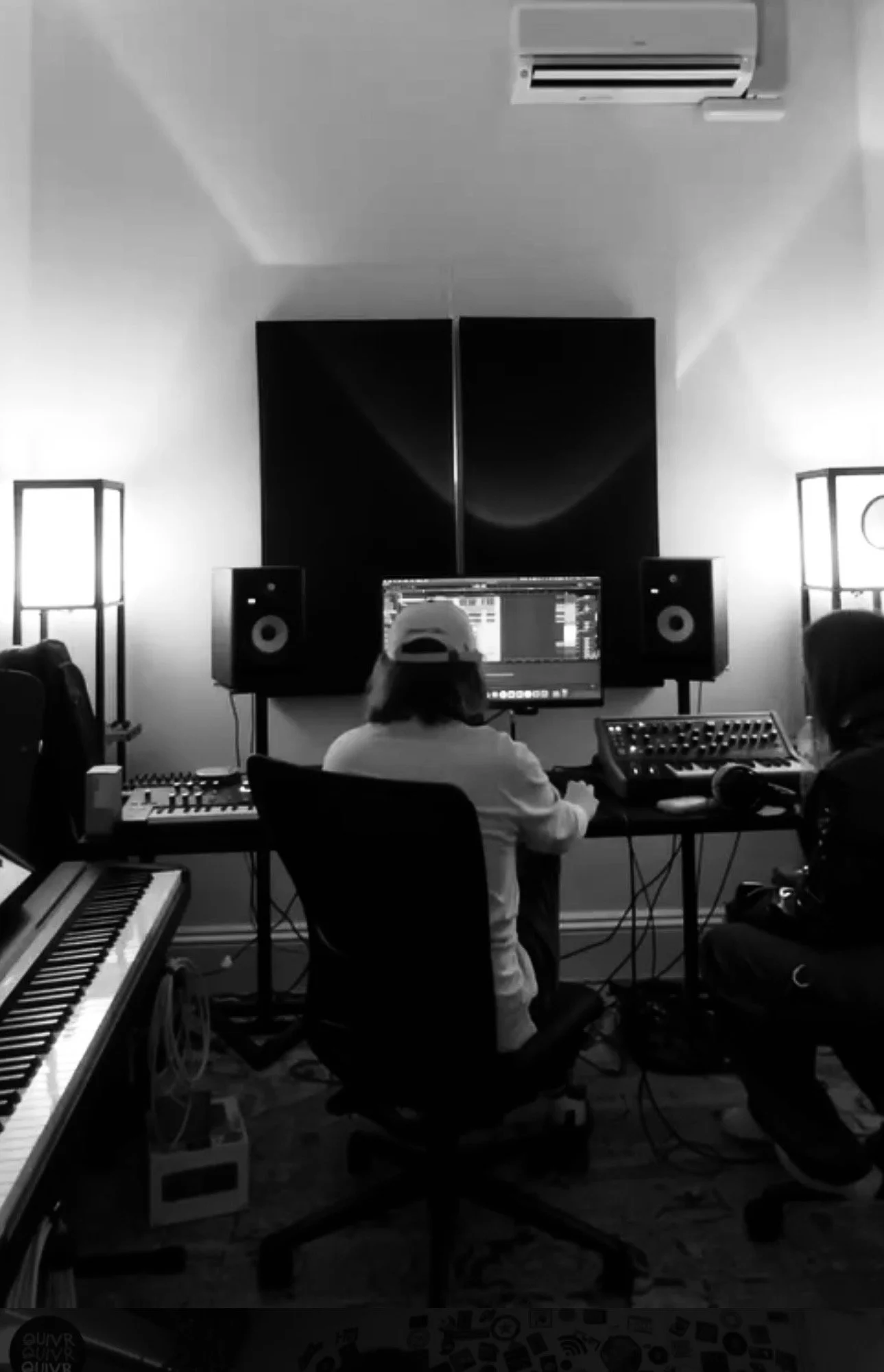 A home music studio with a person working on a computer, surrounded by musical equipment and instruments, including a keyboard, speakers, and a sound mixer.