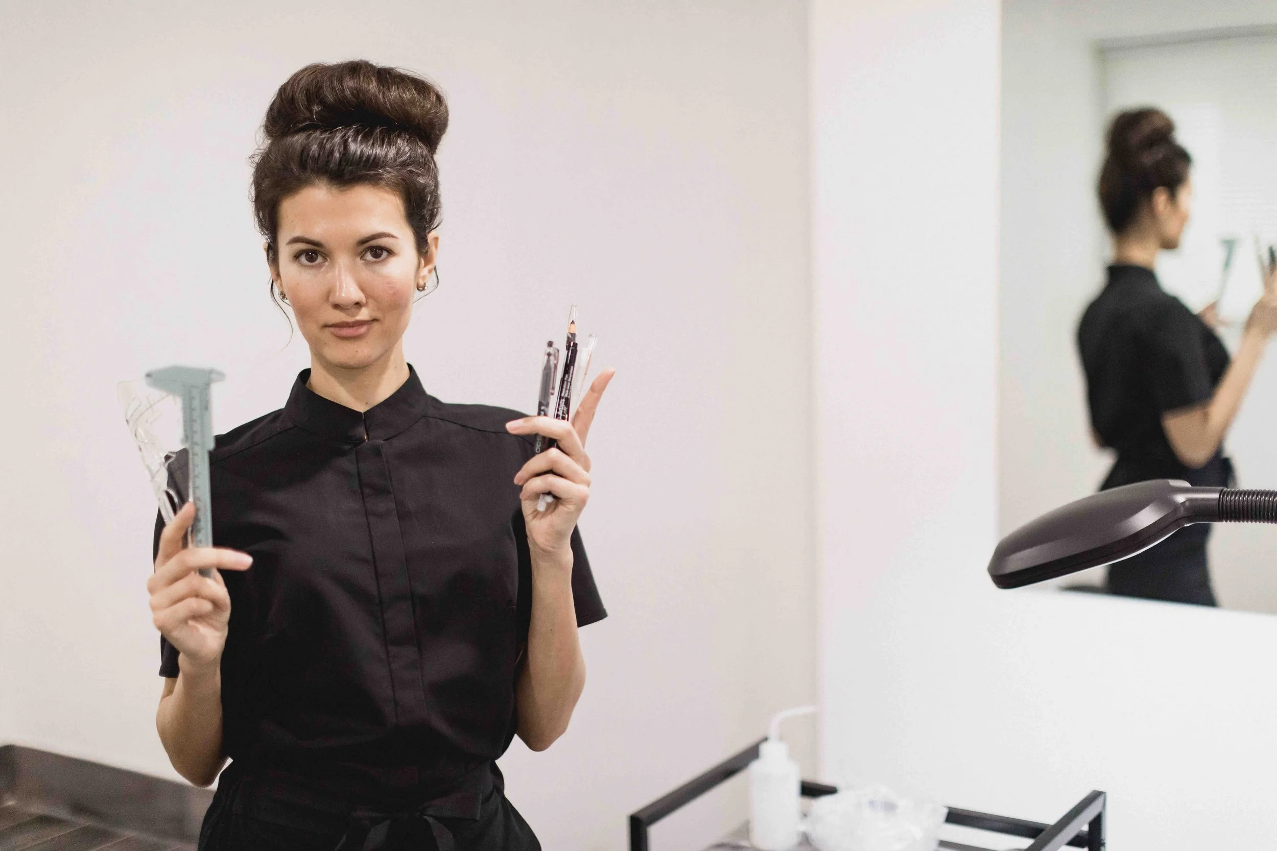 Picture of Karina the permanent makeup artist holding her tools in both hands. Wearing black uniform and having her hair in a bun. Thee background is light grey.