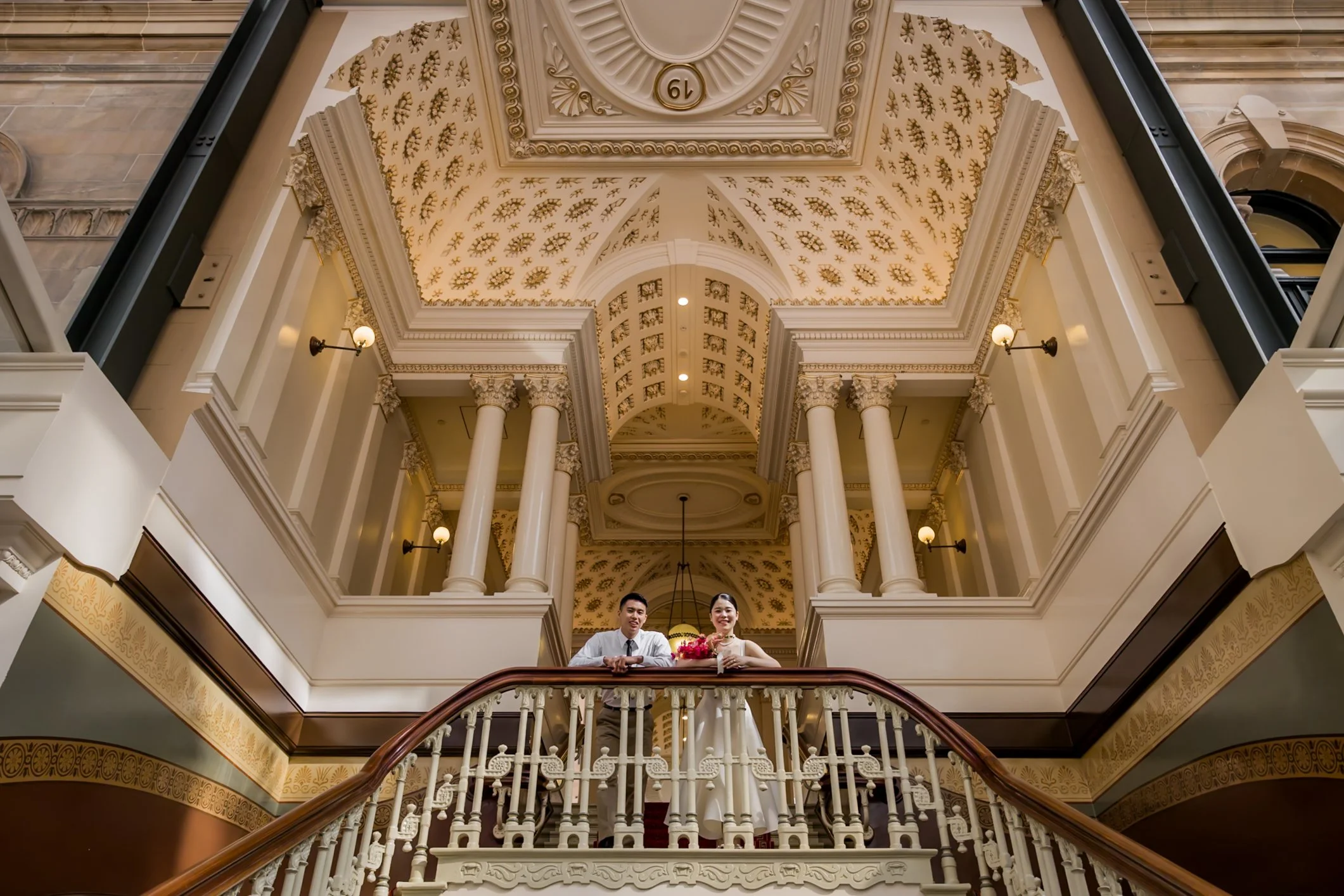 A couple in wedding attire standing on a staircase in an architecturally ornate interior with tall columns, decorative ceiling, and elegant lighting.