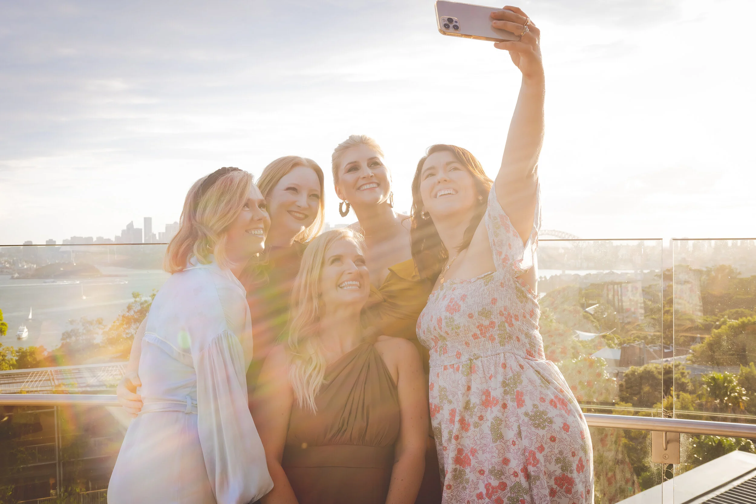 Group of five women taking a selfie on a balcony with a cityscape and river in the background, sunlight creating a lens flare.