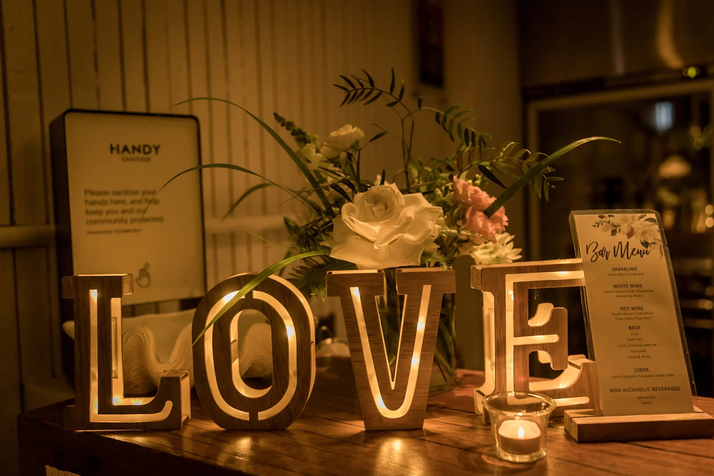 Illuminated wooden letters spell "LOVE" on a candlelit table, with a floral arrangement and a bar menu in the background.