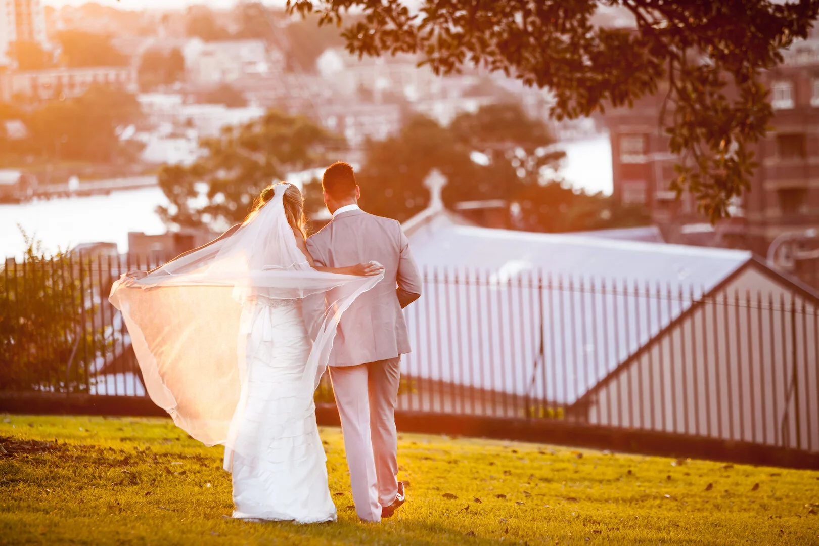 A bride and groom walk together outside during sunset, with the bride's veil flowing behind. They are dressed in wedding attire, with the bride in a white gown and the groom in a light-colored suit. There are buildings and trees in the background.