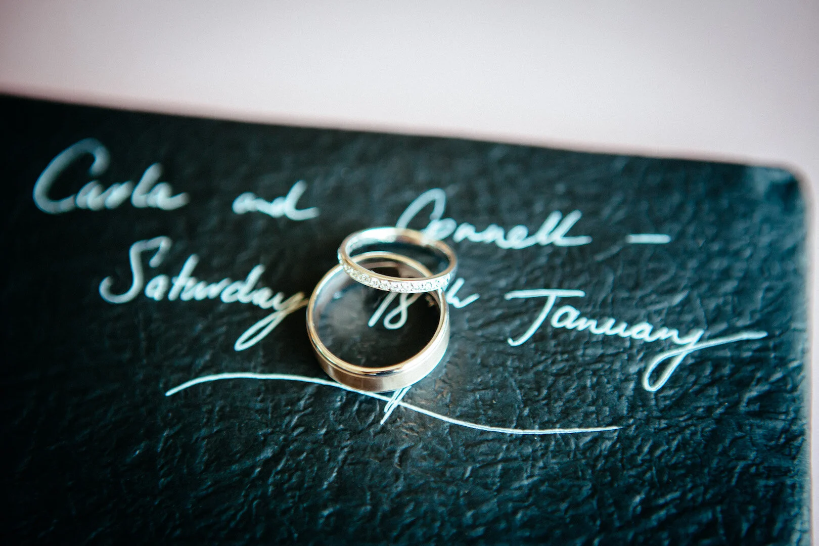 Two wedding rings, one with embedded diamonds, resting on a black textured surface with handwritten white script text.