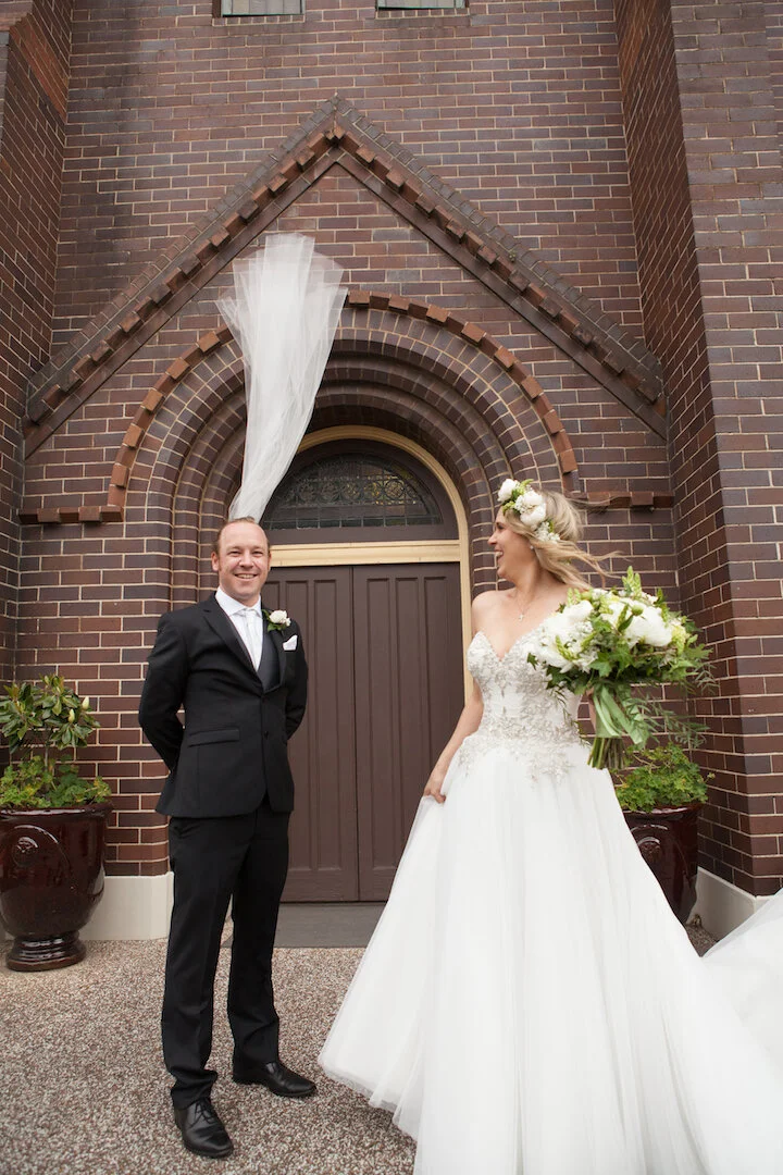 A newlywed couple stands outside a church. The bride holds a bouquet of white flowers with greenery, and the groom is dressed in a black suit, white shirt, and tie. They are smiling and looking at each other, with a brick church building and a doorwa