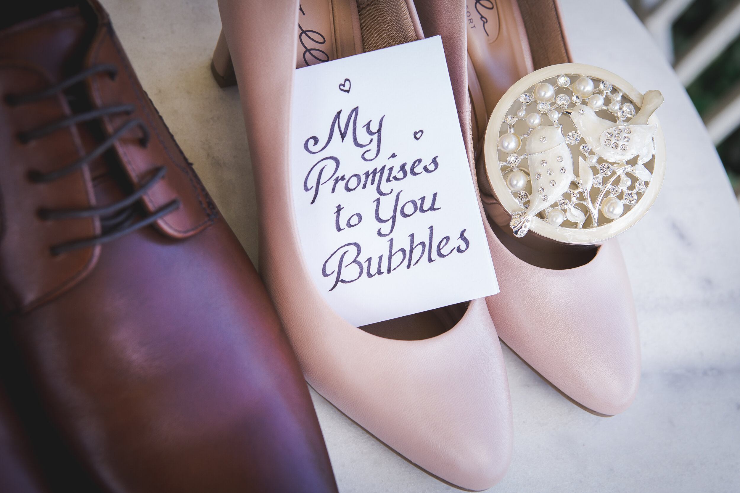 A pair of pink high-heeled shoes, a note with the handwritten phrase 'My Promises to You Bubbles,' a large brown leather shoe, and a decorative round container filled with pearls, rhinestones, and small ornamental birds.