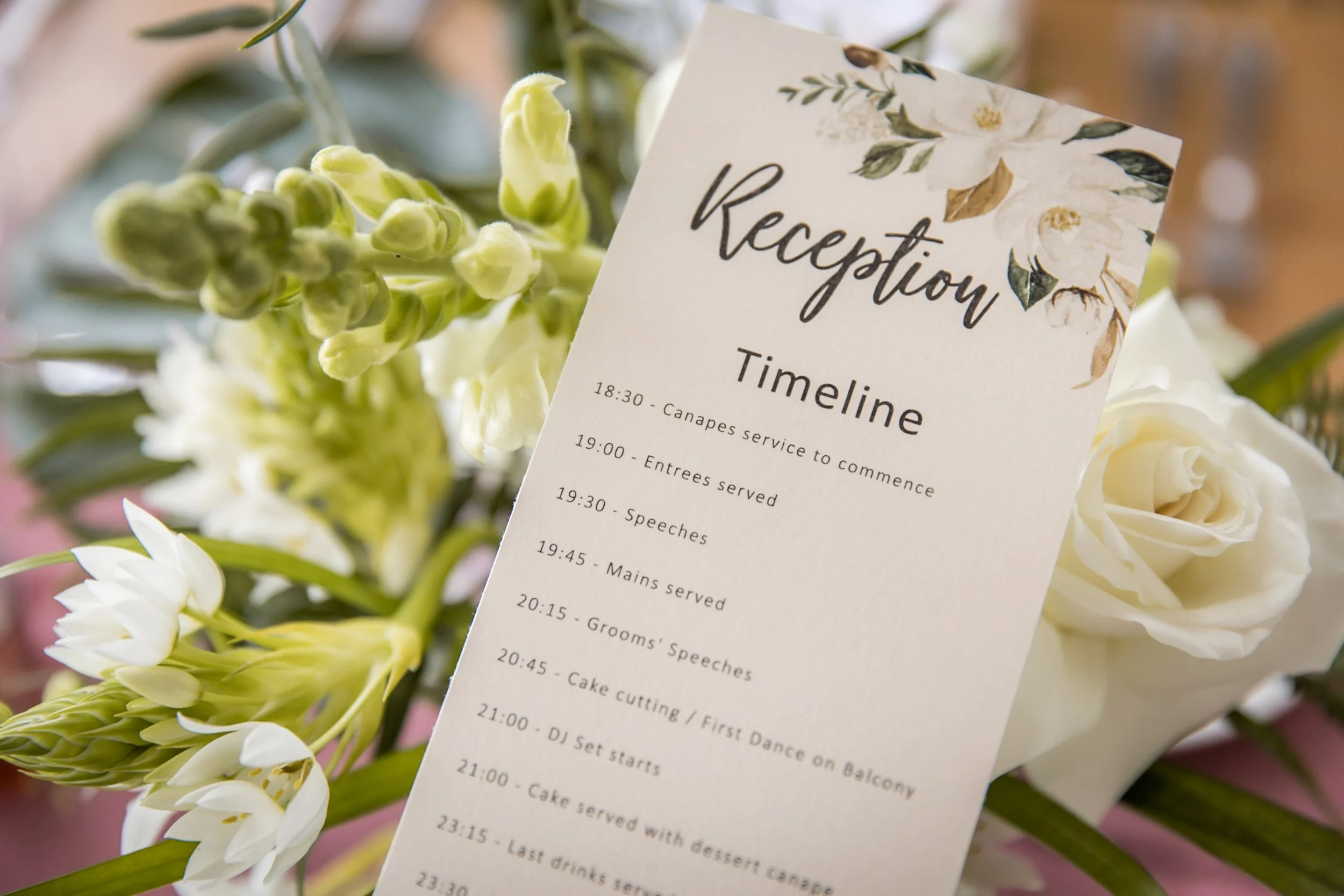 Reception event schedule printed on a piece of paper, placed against floral decorations including white roses and other white and green flowers.