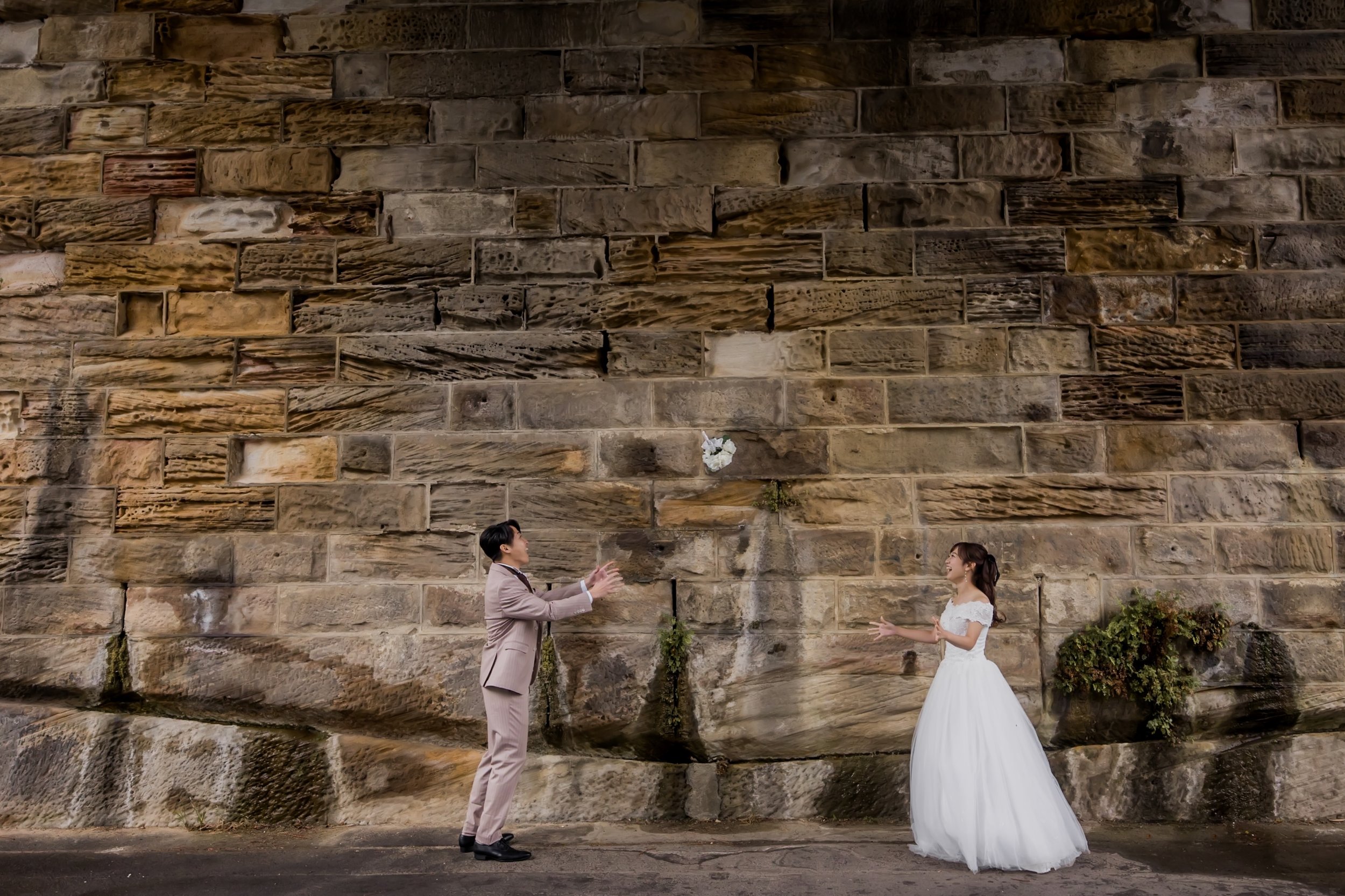 A man and woman in wedding attire are standing in front of a large stone wall, with the man throwing a bouquet of flowers toward the woman, who is smiling with her arms open.