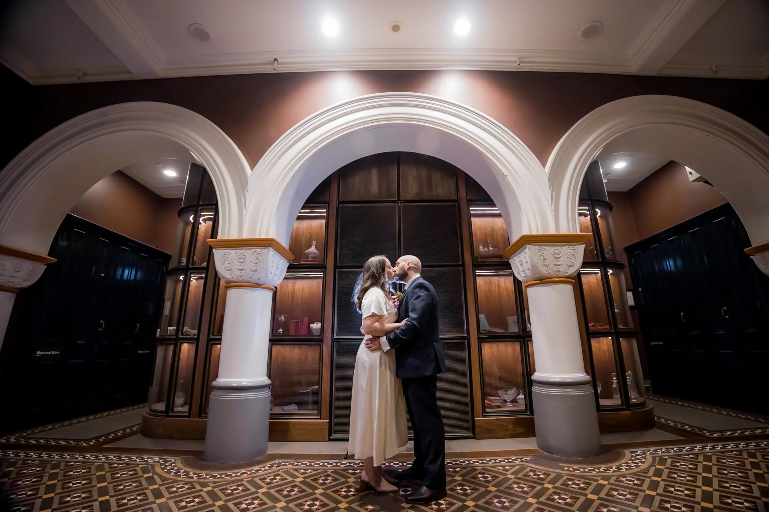 A couple sharing a kiss in formal attire inside an ornate room with decorative columns, wooden cabinets, and patterned flooring. - The Tea Room QVB
