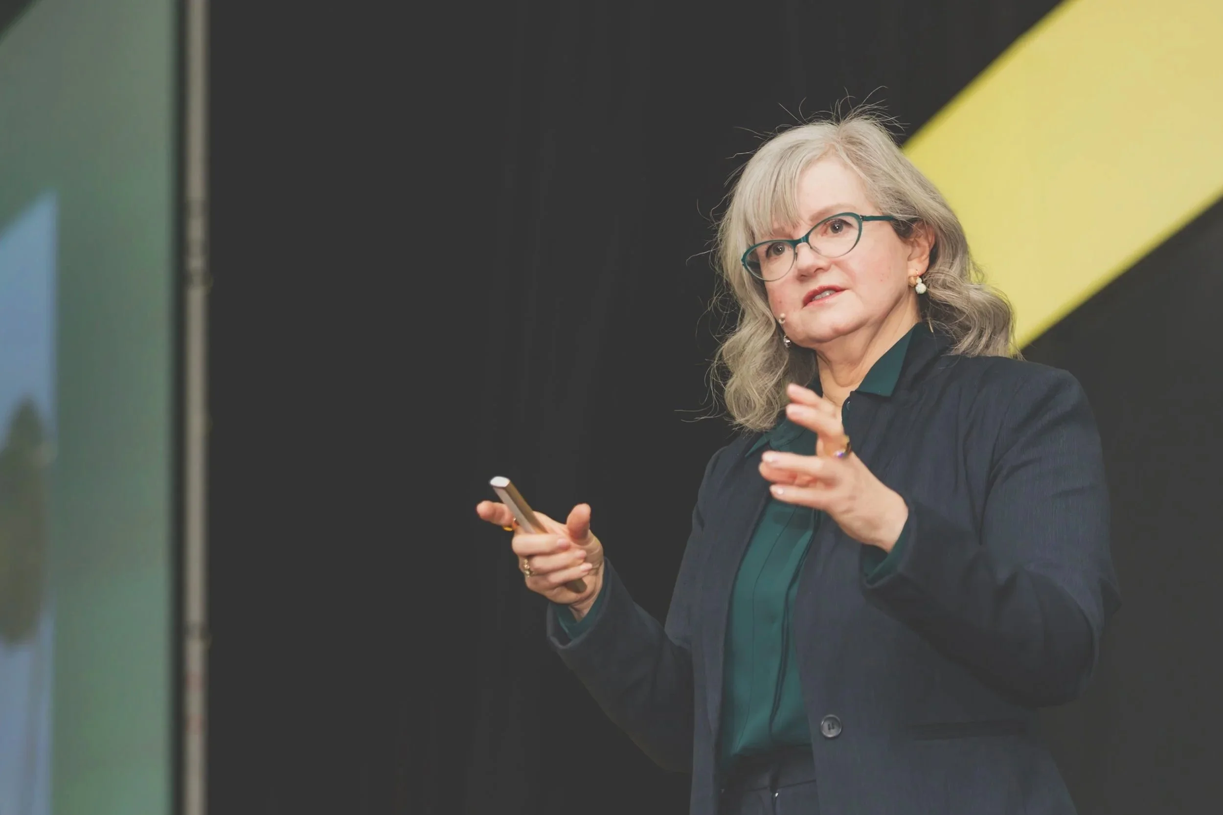 A woman with gray hair, glasses, and pearl earrings giving a presentation or speech on stage, holding a wireless clicker in her right hand, in a professional setting.