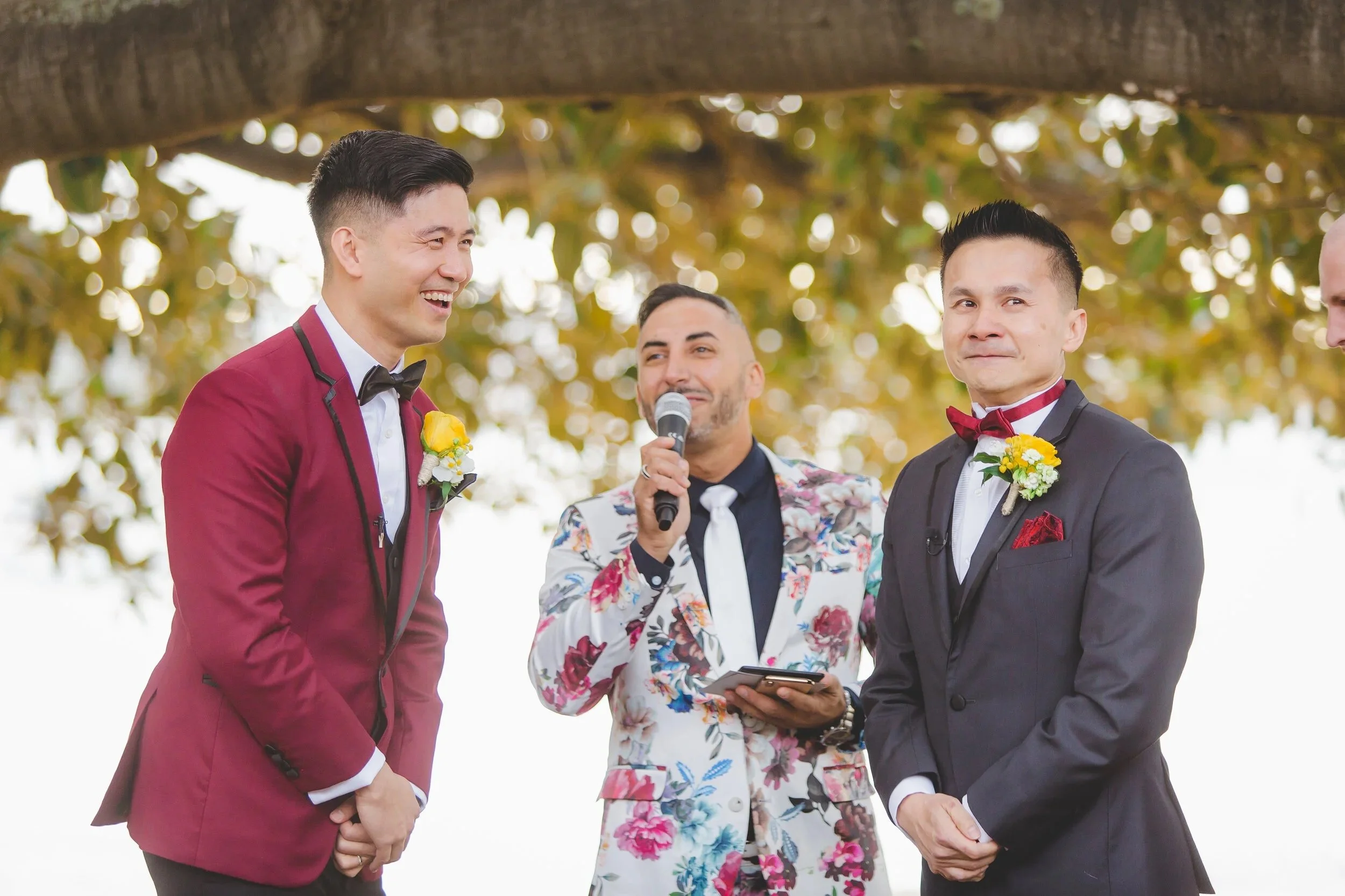 Two men in suits and a person in floral blazer standing outdoors during a wedding ceremony, with leaf-covered trees in the background.