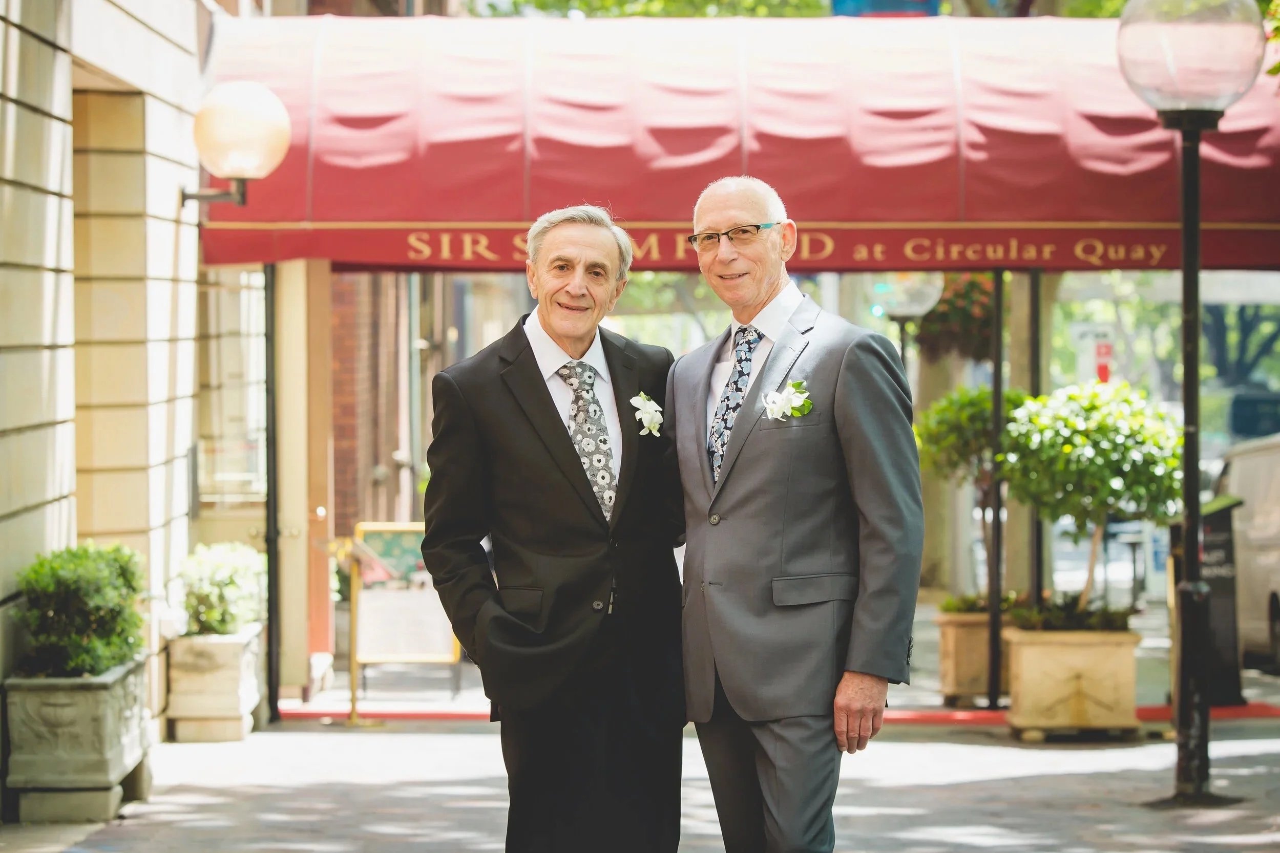 Two men in suits with boutonnières standing on a city sidewalk in front of a restaurant entrance, smiling at the camera.