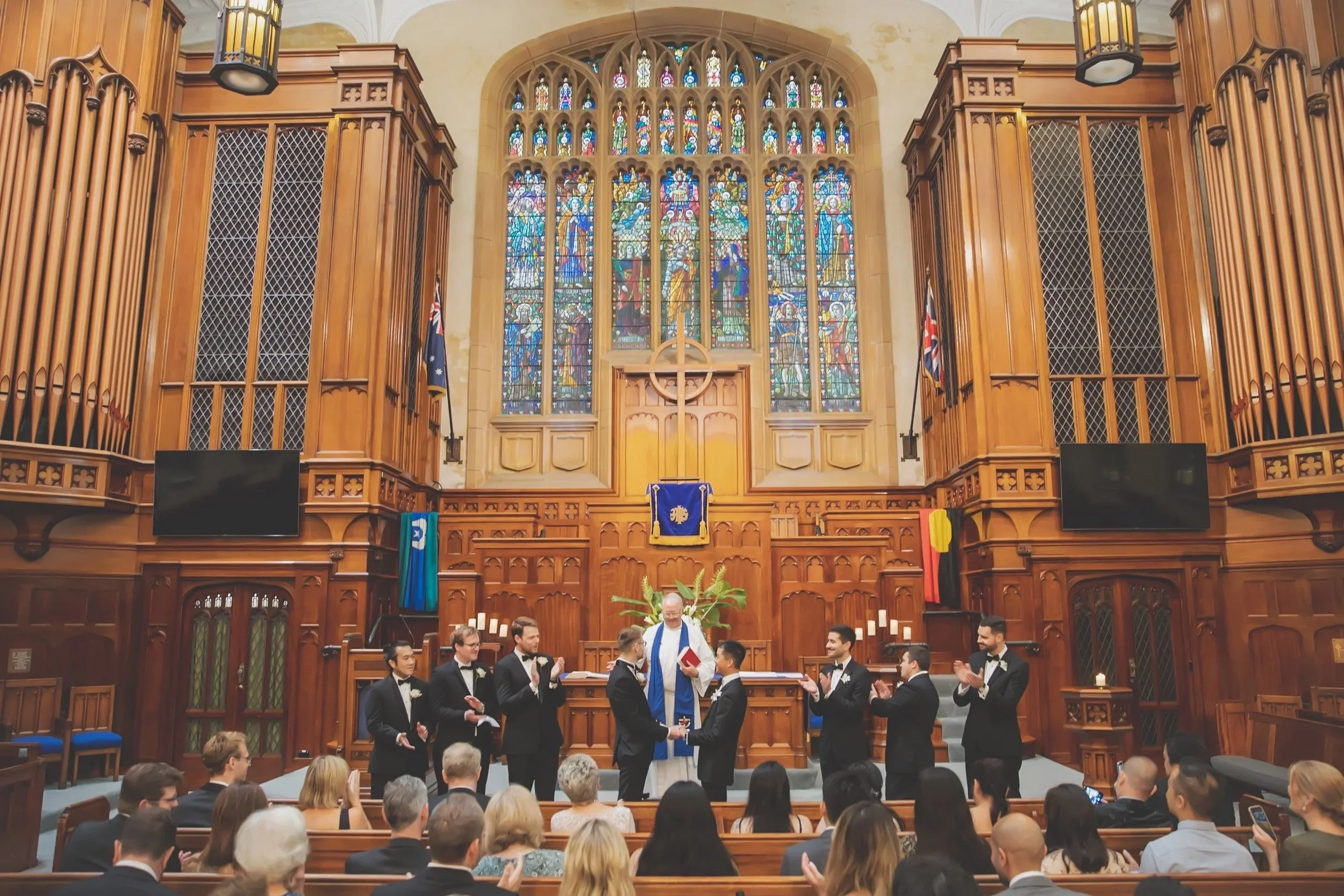 A wedding ceremony inside a church with stained glass windows depicting religious figures. The couple is exchanging vows at the altar, surrounded by friends and family. The church has wooden paneling and flags on either side.