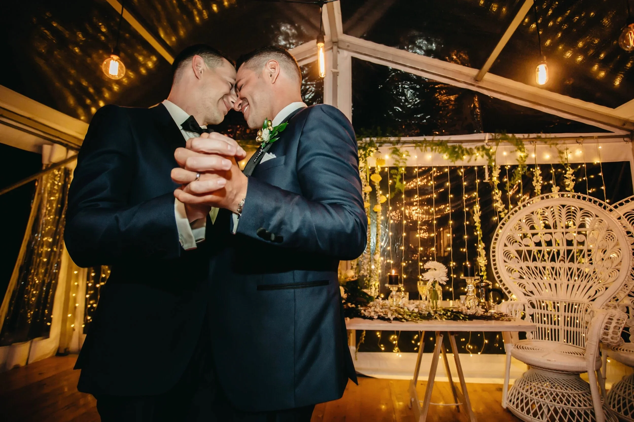 Two men in tuxedos sharing a dance at their wedding reception under string lights and decorated with flowers and candles.