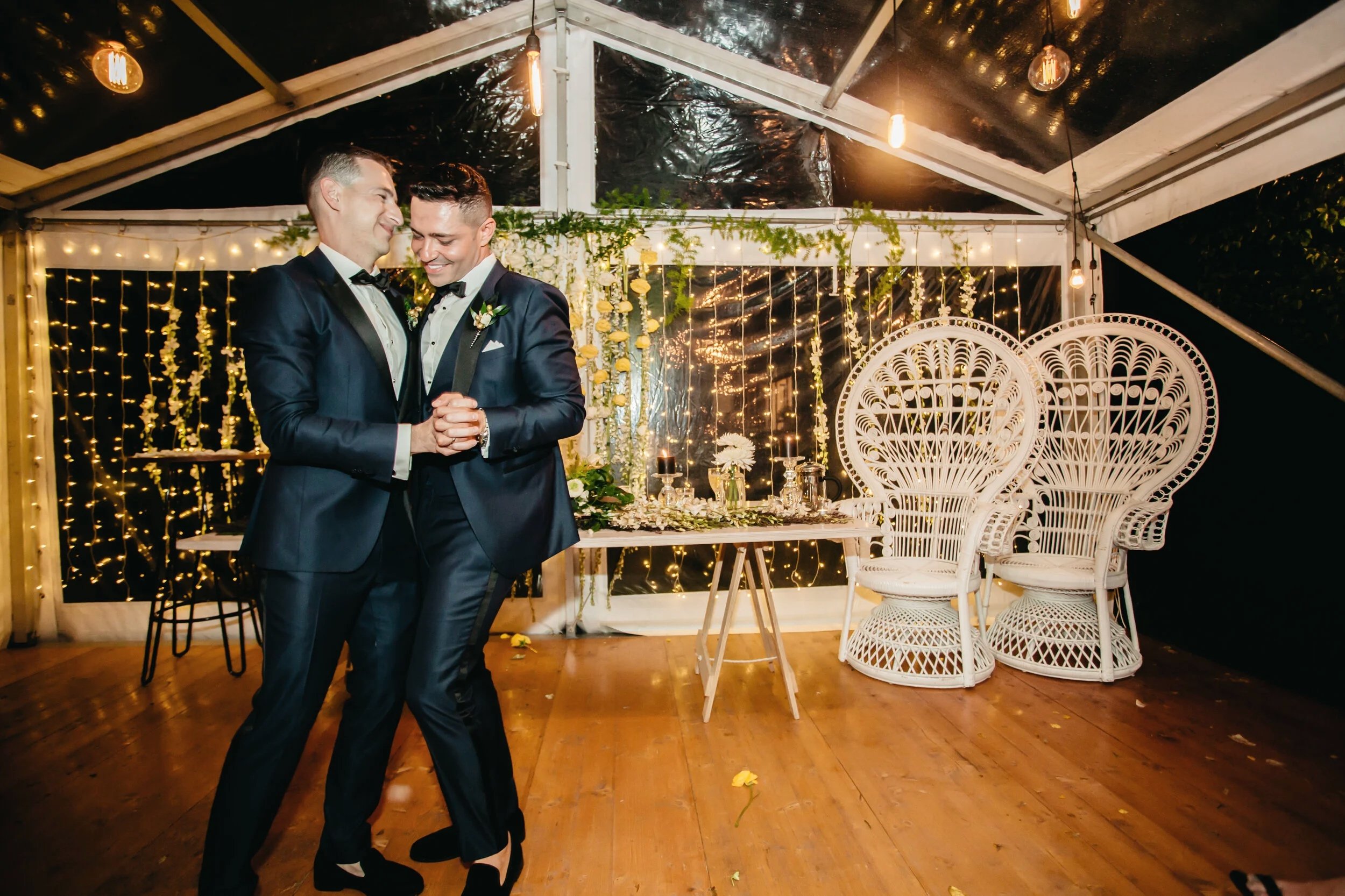 Two men in tuxedos dance together in a decorated outdoor wedding tent with string lights and white wicker chairs.