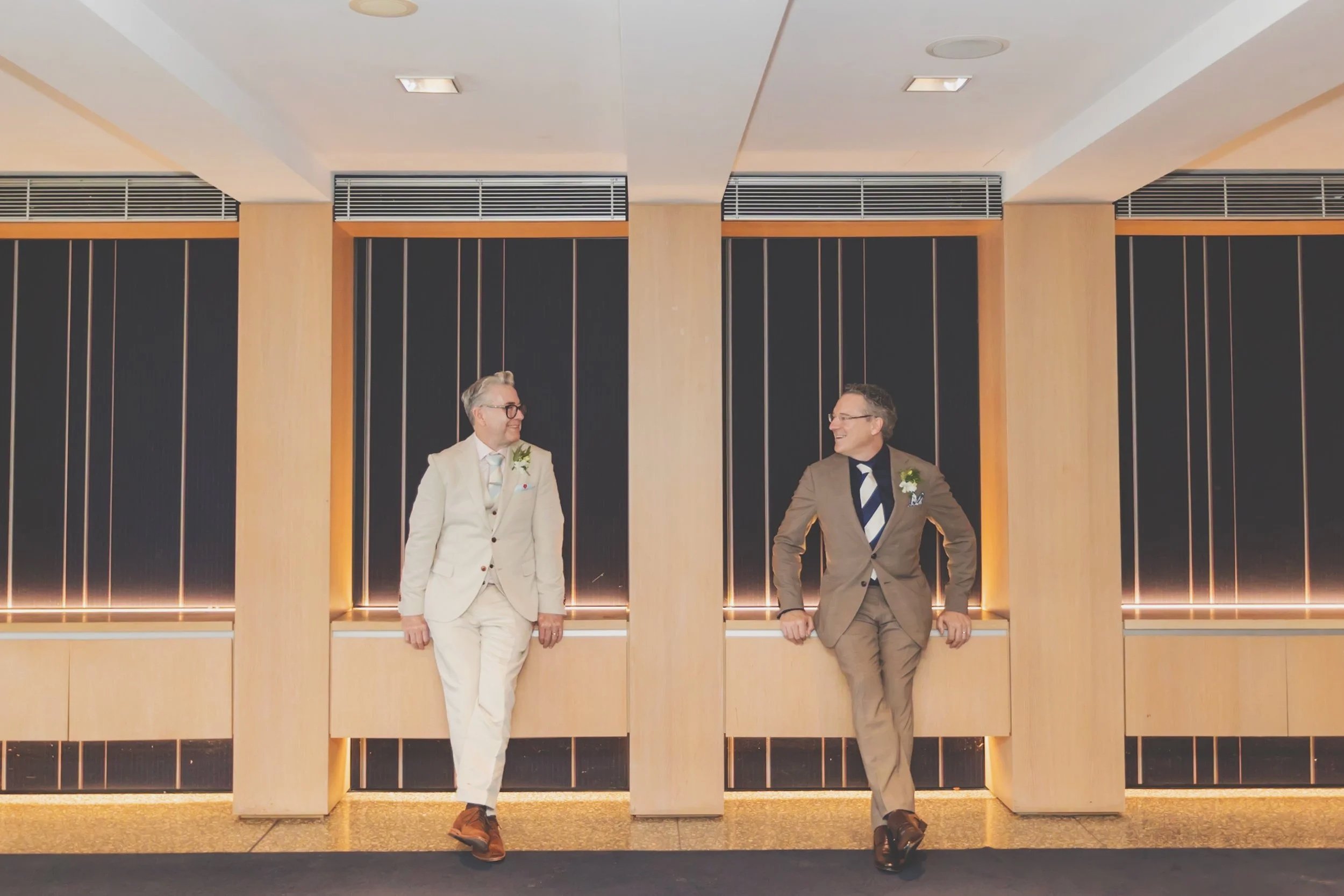 Two men in suits standing in a modern hallway, smiling and looking at each other.