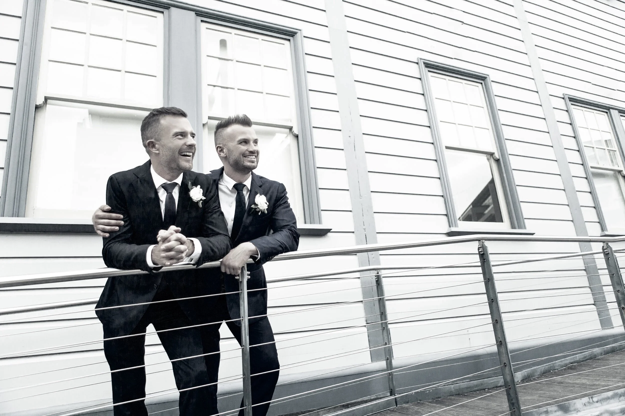 Two men in blue suits with white shirts and ties, wearing white boutonnieres, standing on a balcony outside a house, smiling and looking into the distance.