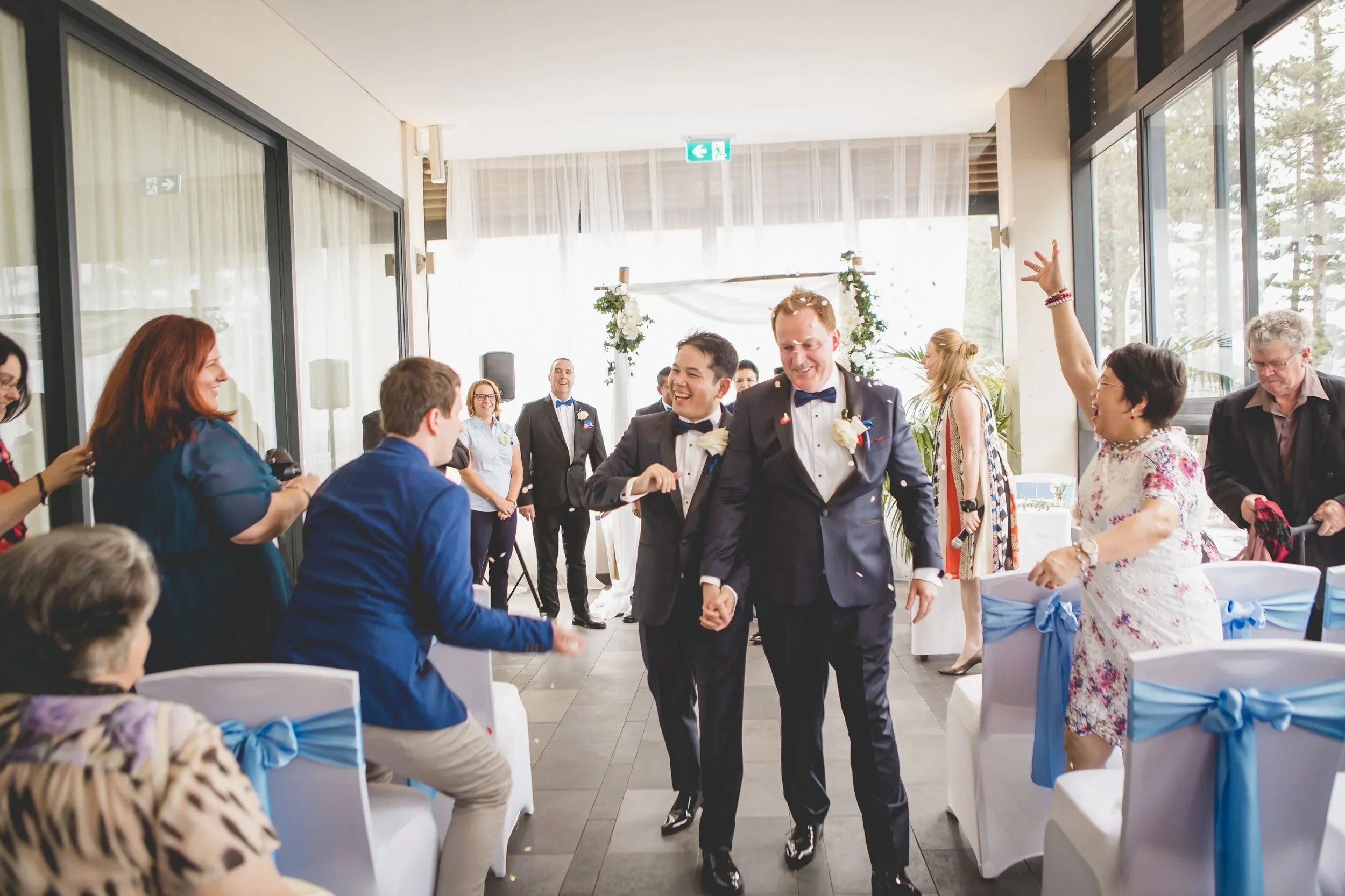 Two men in tuxedos holding hands and smiling as they walk through a celebration, surrounded by people cheering and throwing confetti at a wedding reception.