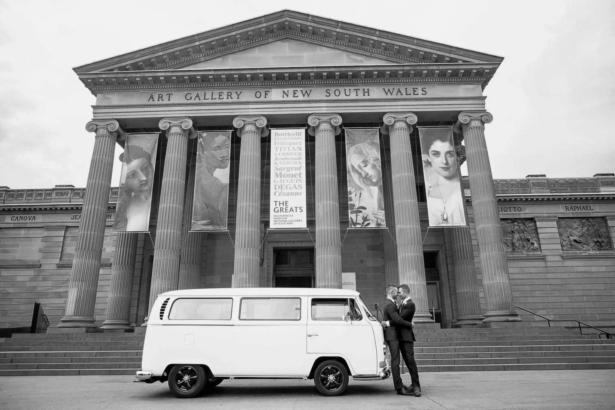 Two men in suits standing close together outside the Art Gallery of New South Wales, a large building with classical architecture and tall columns, with banners displaying artwork. A white vintage van is parked nearby.