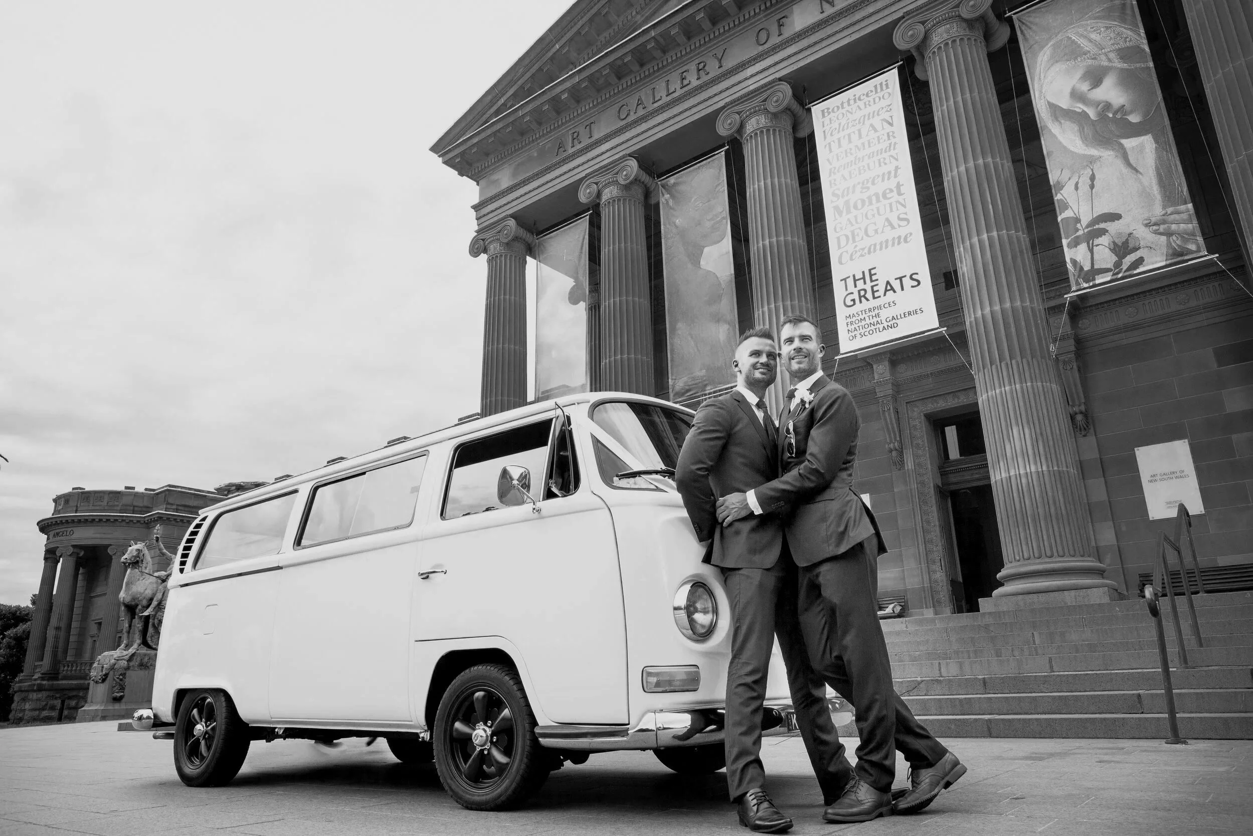 Two men in suits holding hands and smiling in front of a white vintage van outside a historic art gallery building. The building has large columns and banners advertising art exhibitions.