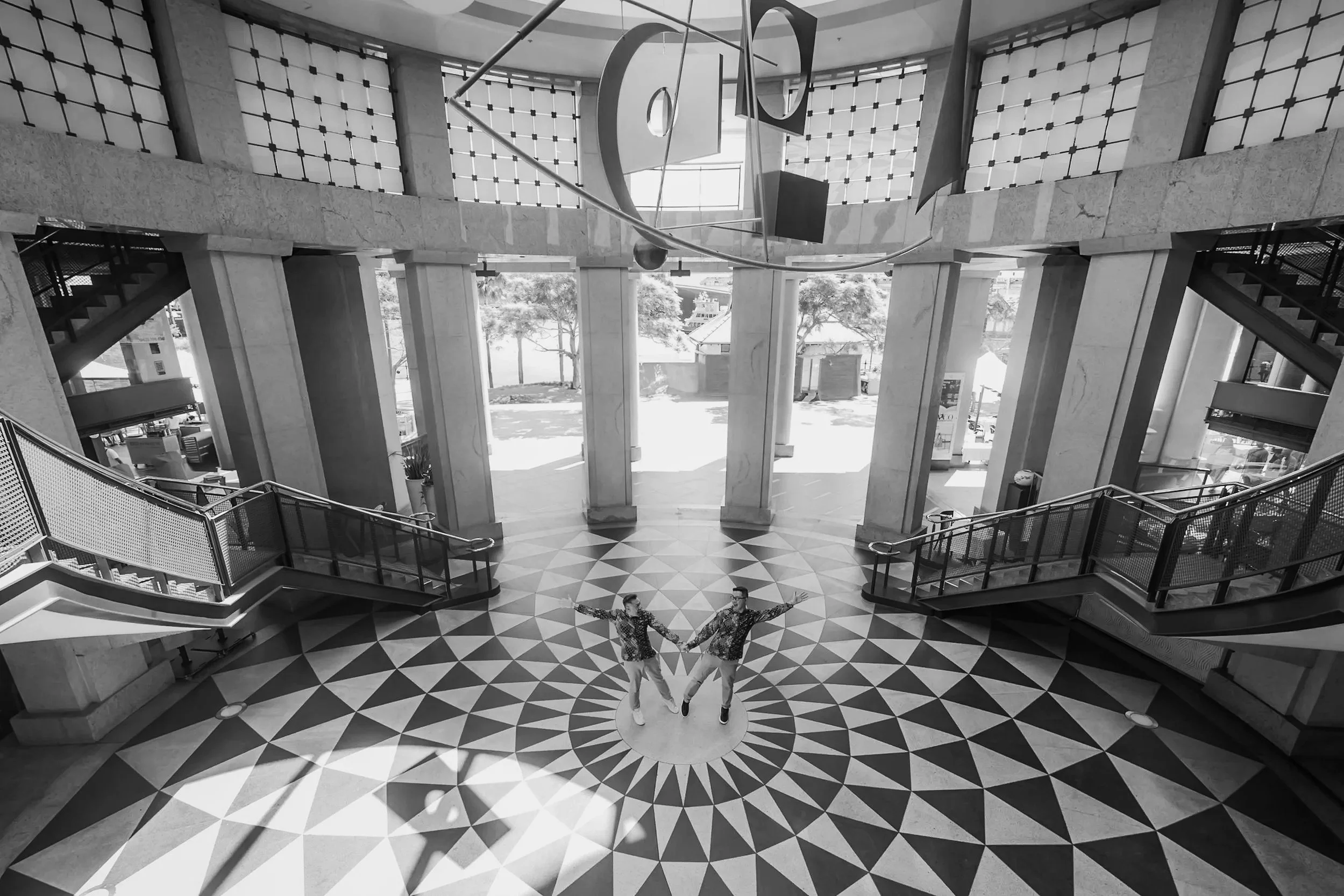 Indoor lobby with geometric black and white marble floor, large windows, modern stairs, and two men holding hands and dancing in the center.