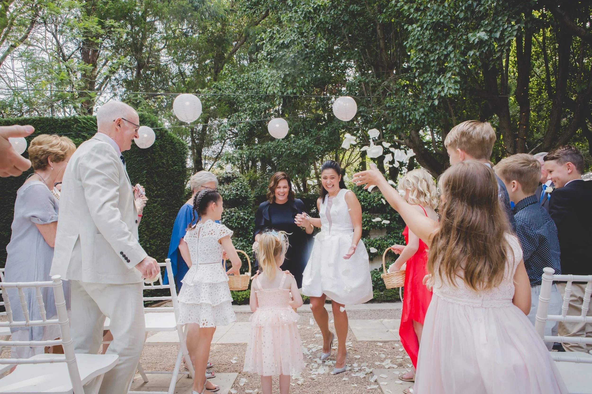Wedding celebration outdoors with guests throwing flower petals, a bride in a white dress smiling, surrounded by children and adults in formal attire, decorated with paper lanterns hanging in the trees.