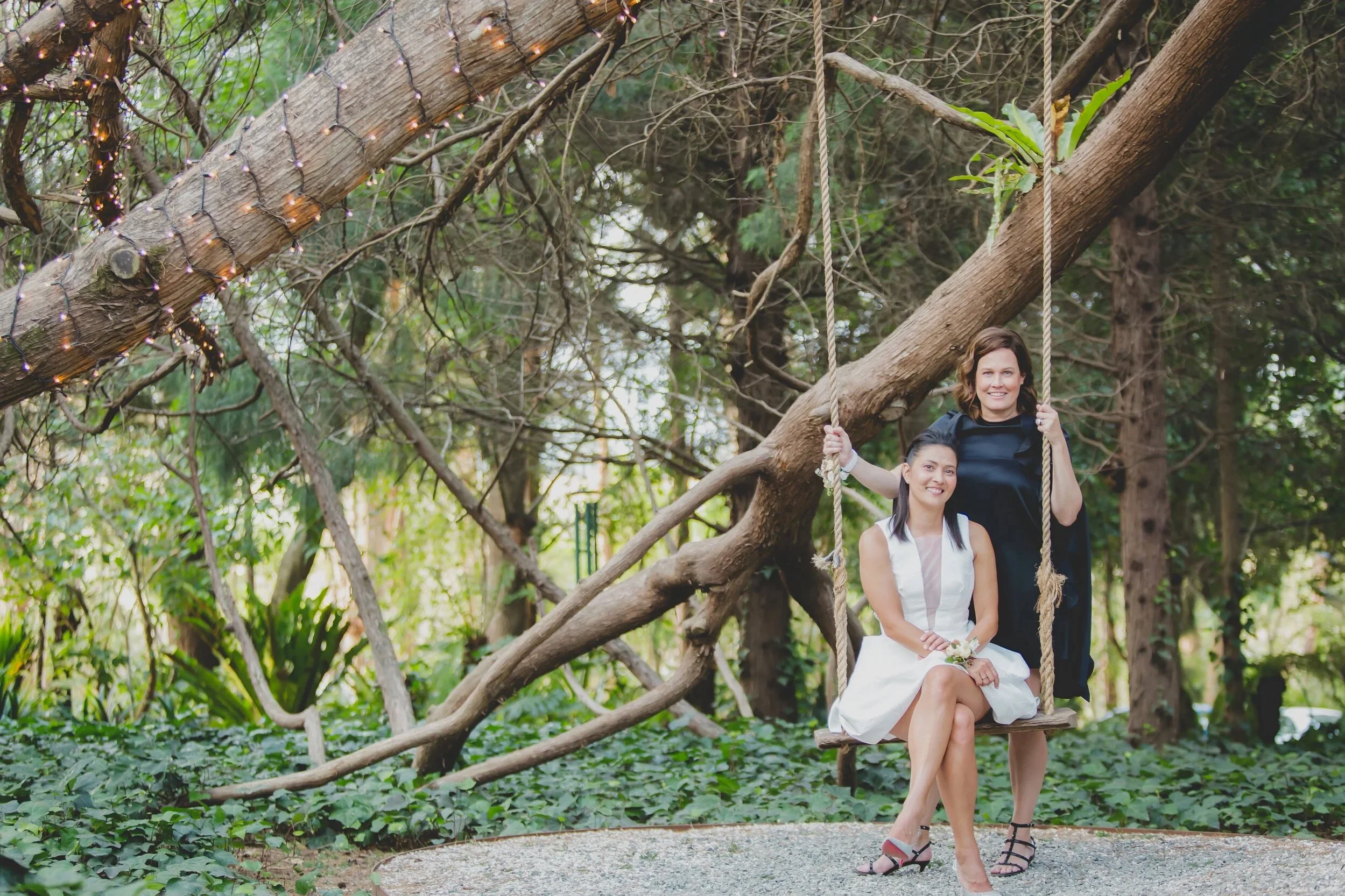 Two women posing on a wooden swing hanging from a tree in a park, with one woman sitting and the other standing behind her. The scene is outdoors with greenery and fairy lights wrapped around the trees.