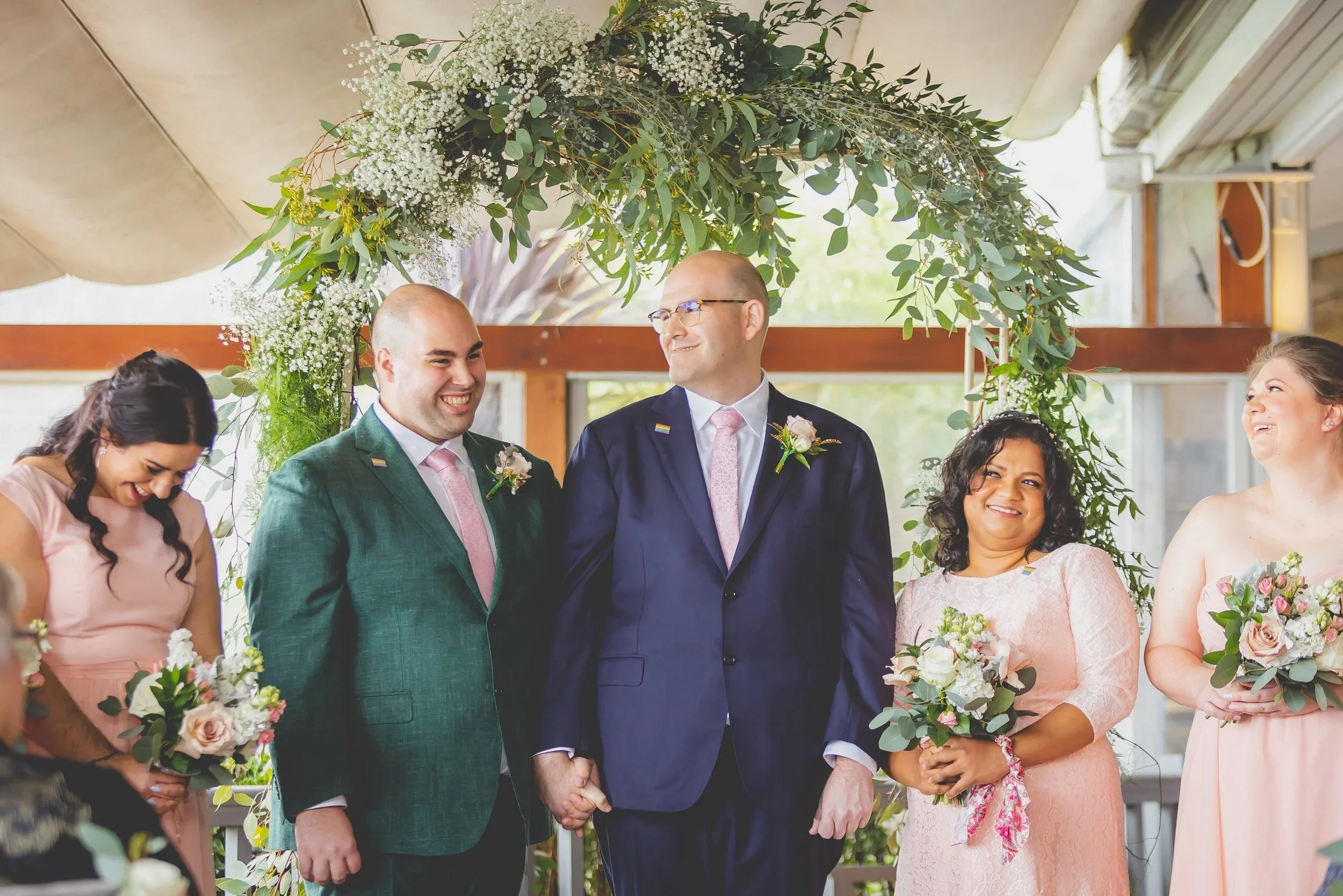 A group of people at a wedding ceremony, with two men holding hands in the center, surrounded by women holding bouquets, under a floral arch.