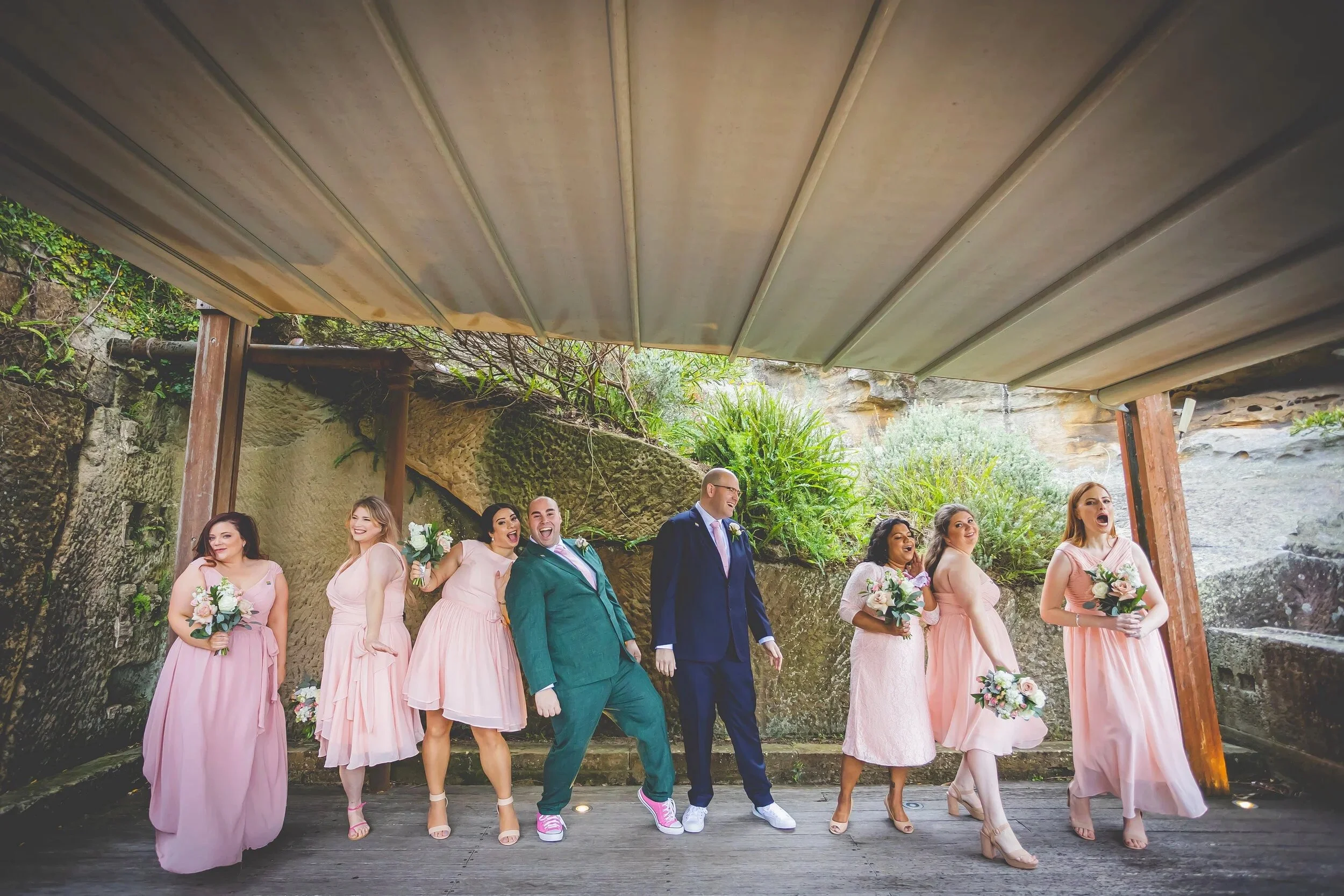 A group of eight people, including bridesmaids and groomsmen, are standing under a large outdoor canopy with greenery and rocks in the background. The women are wearing pink dresses and holding bouquets, while the men are in suits. Everyone appears h