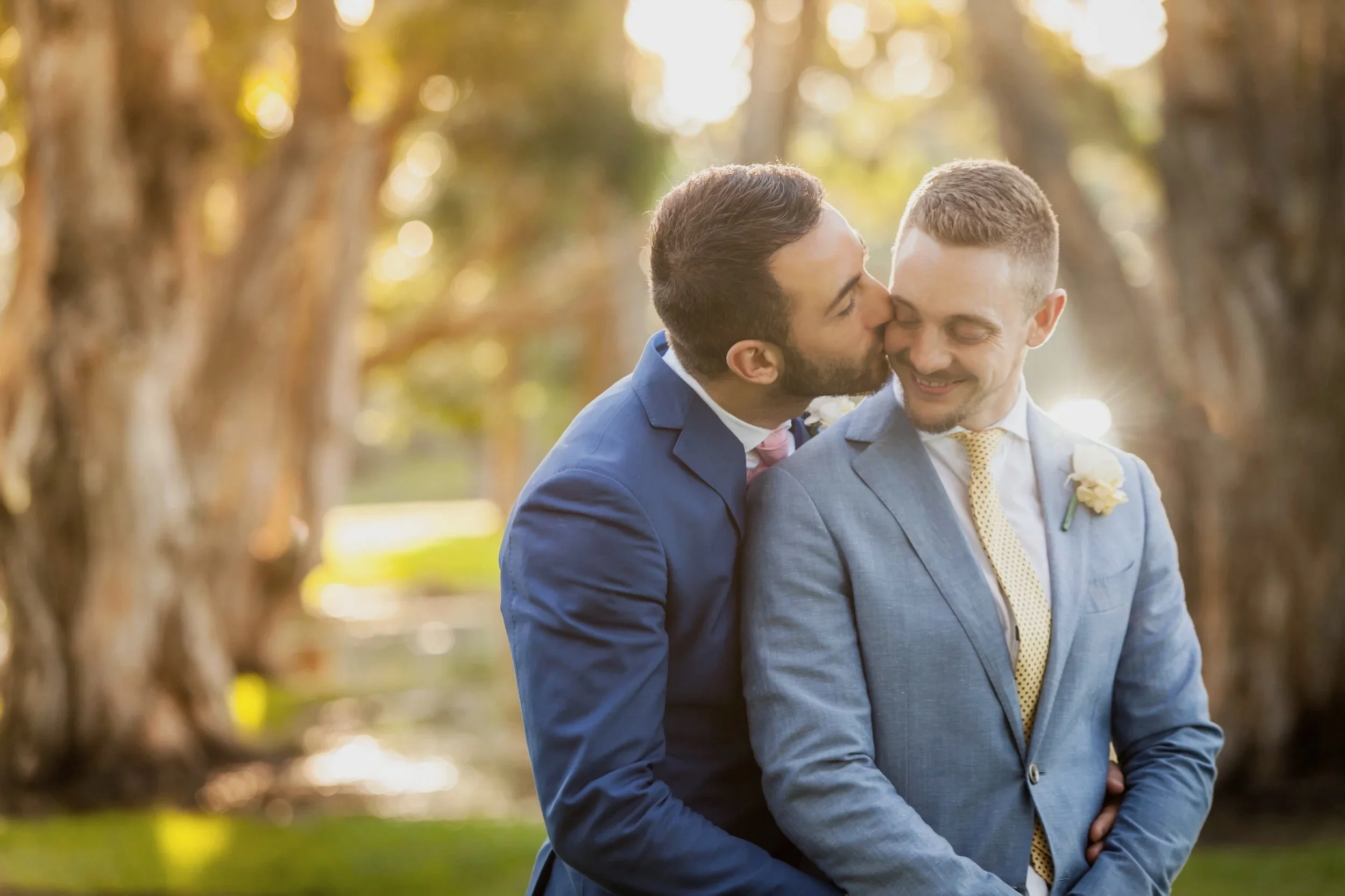 Two men in suits share a kiss at their wedding outdoors during sunset, with trees and warm light in the background.