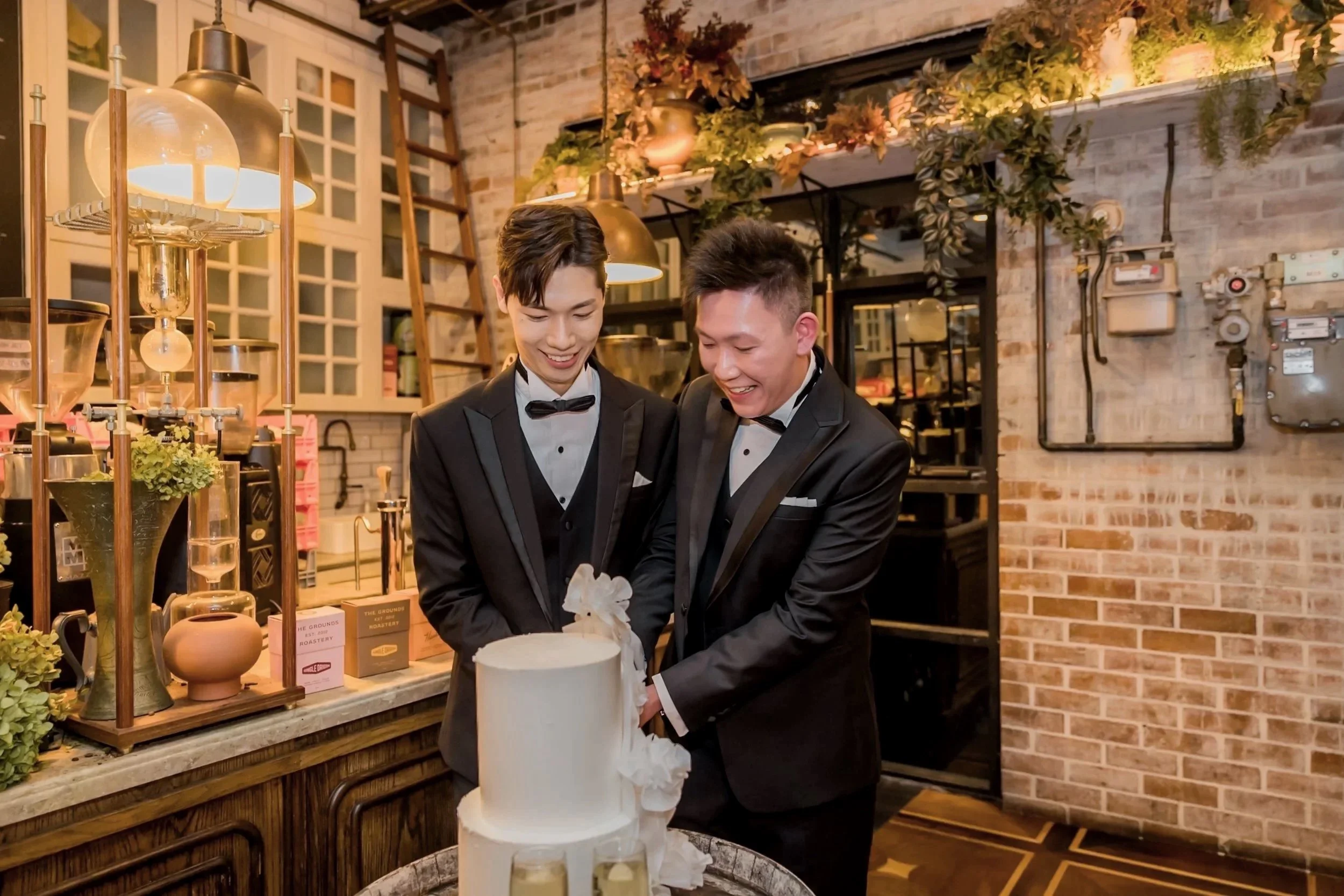 Two men in black tuxedos cutting a wedding cake in a decorated restaurant or cafe with brick walls and warm lighting.