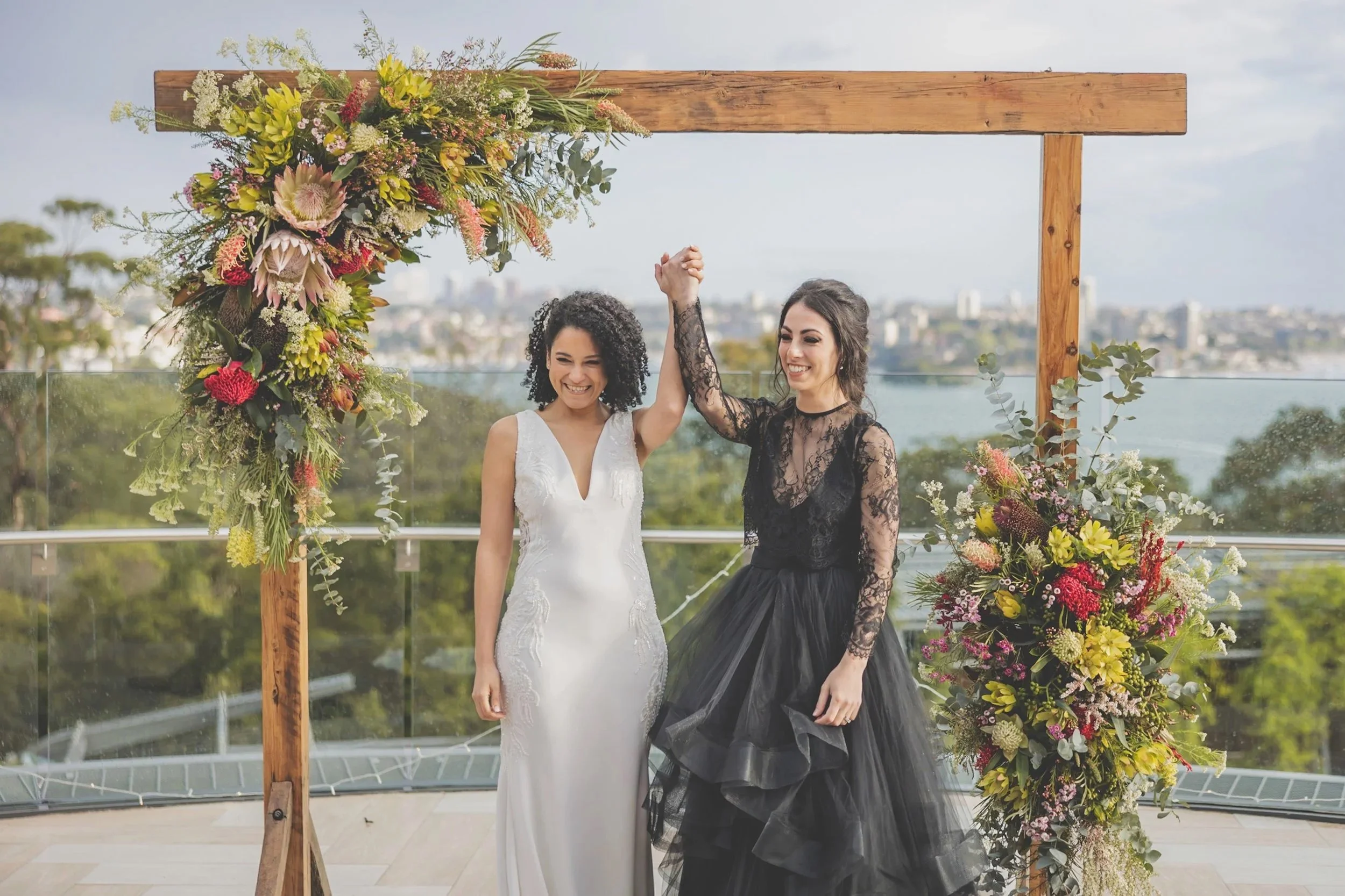 Two women, one in a white wedding dress and the other in a black dress with lace, celebrating under a floral wedding arch with a scenic outdoor view in the background.