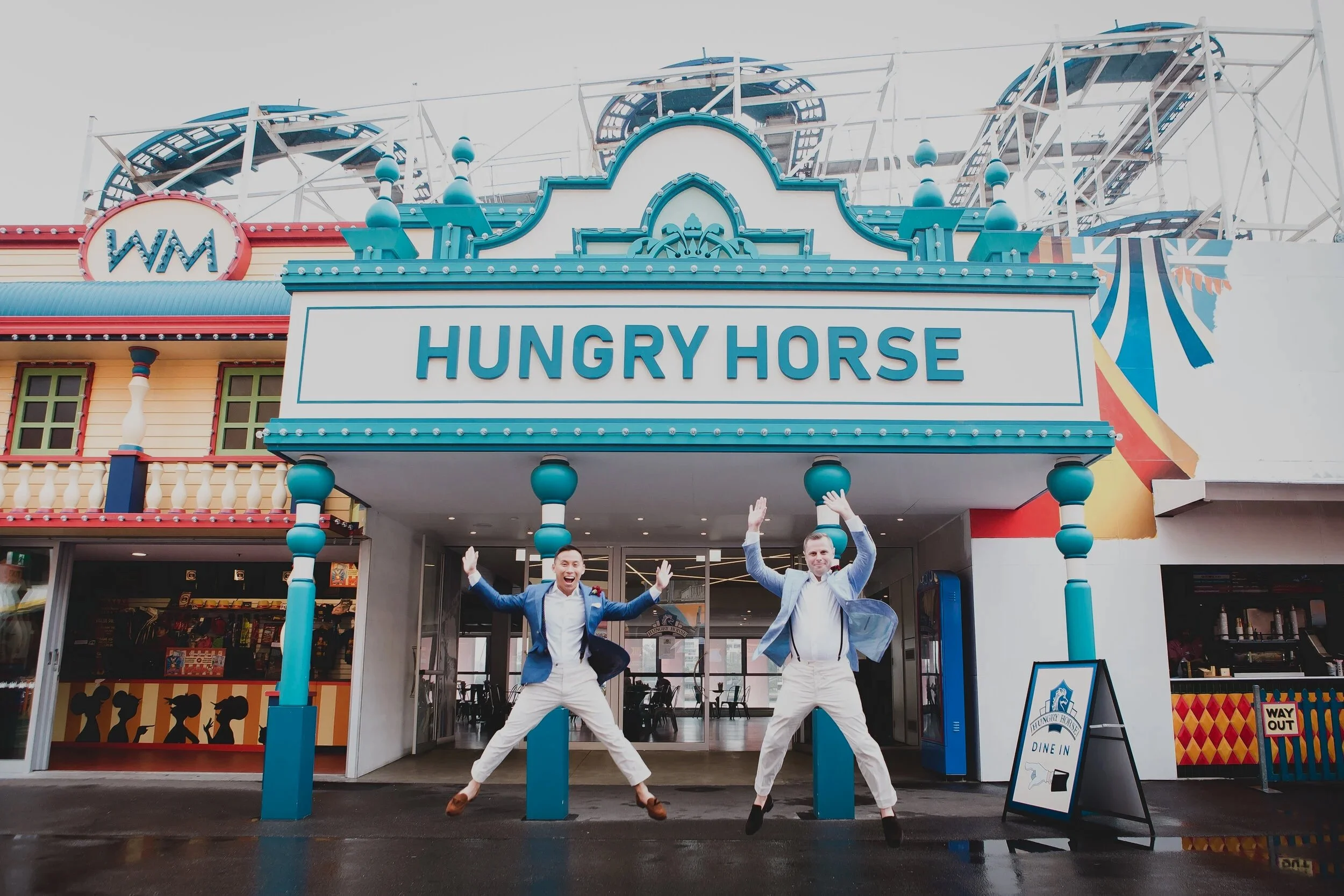 Two men in suits jumping in front of the Hungry Horse amusement park entrance, smiling with arms raised
