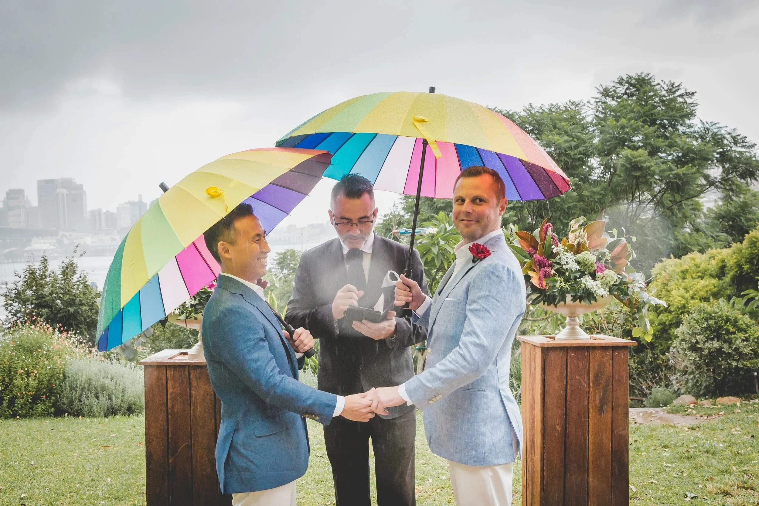 A wedding ceremony outdoors under a multicolored umbrella, with two men holding hands and exchanging vows, officiant reading from a book, and city skyline in the background.