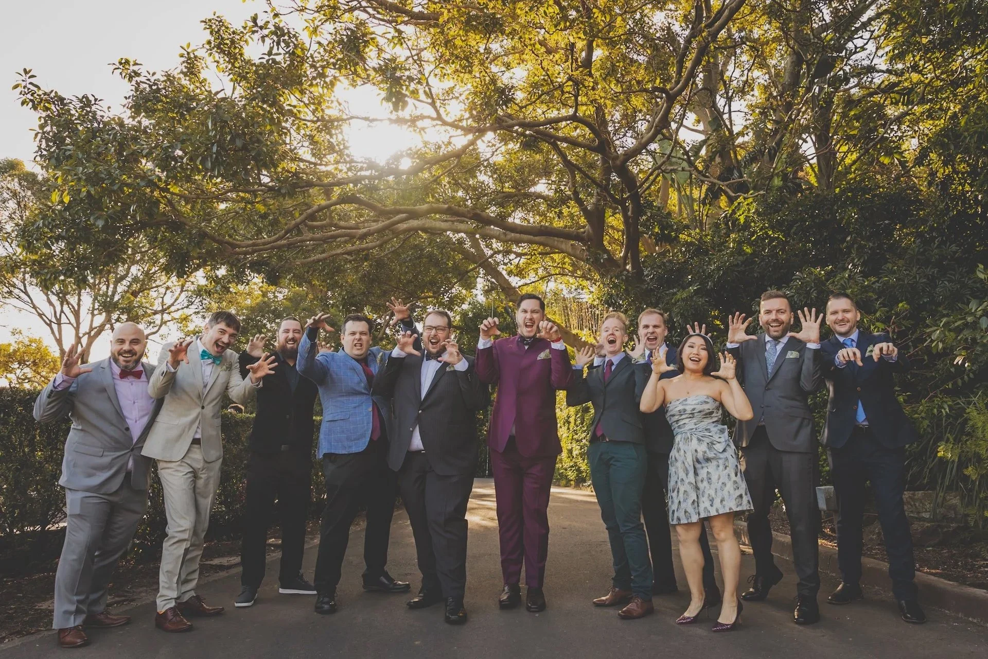 Group of people wearing formal attire, posing playfully outdoors with trees and sunlight in the background.