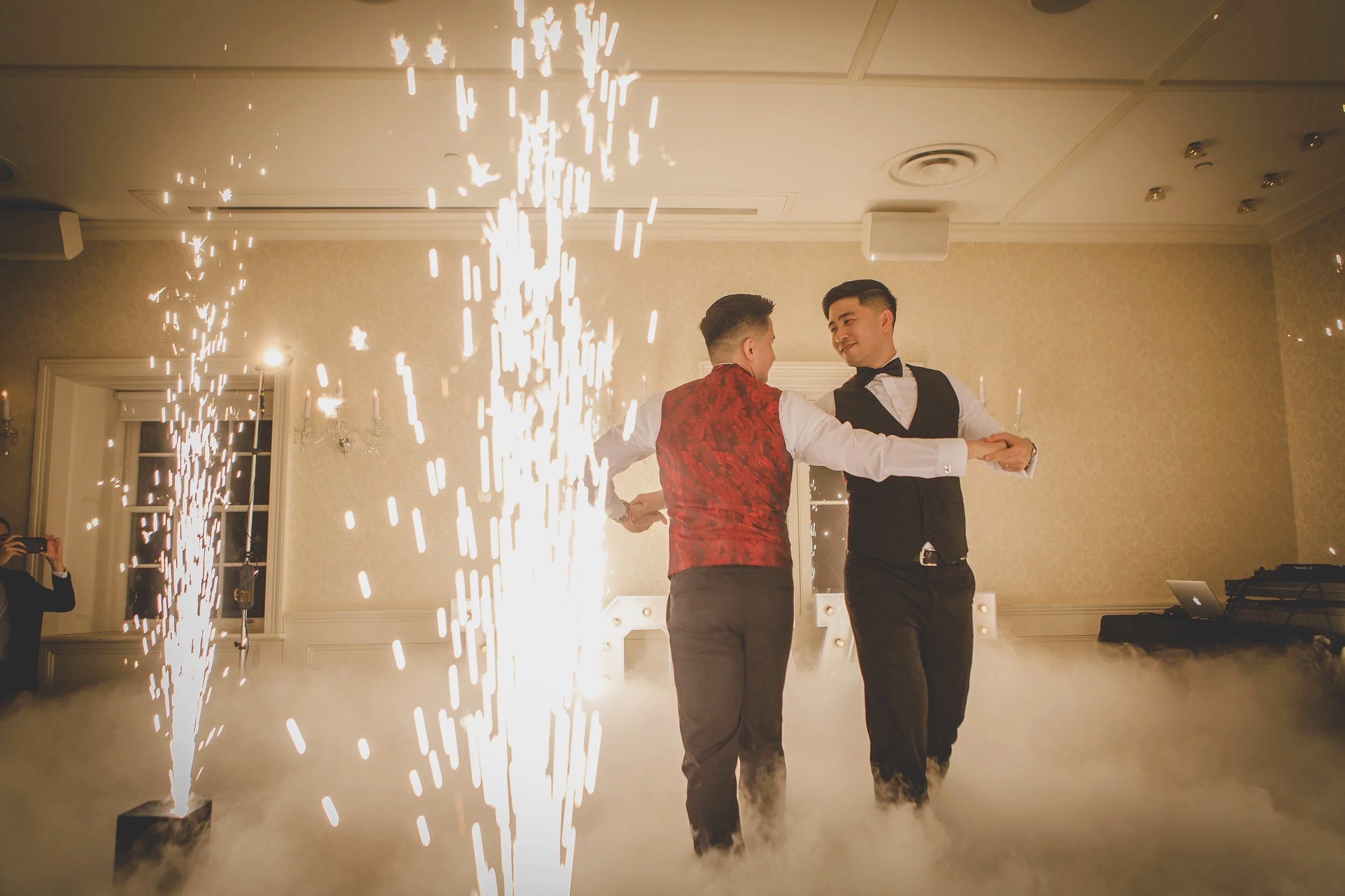 Two men dancing at a celebration with sparks and smoke effects, dressed in formalwear, in a decorated indoor venue.