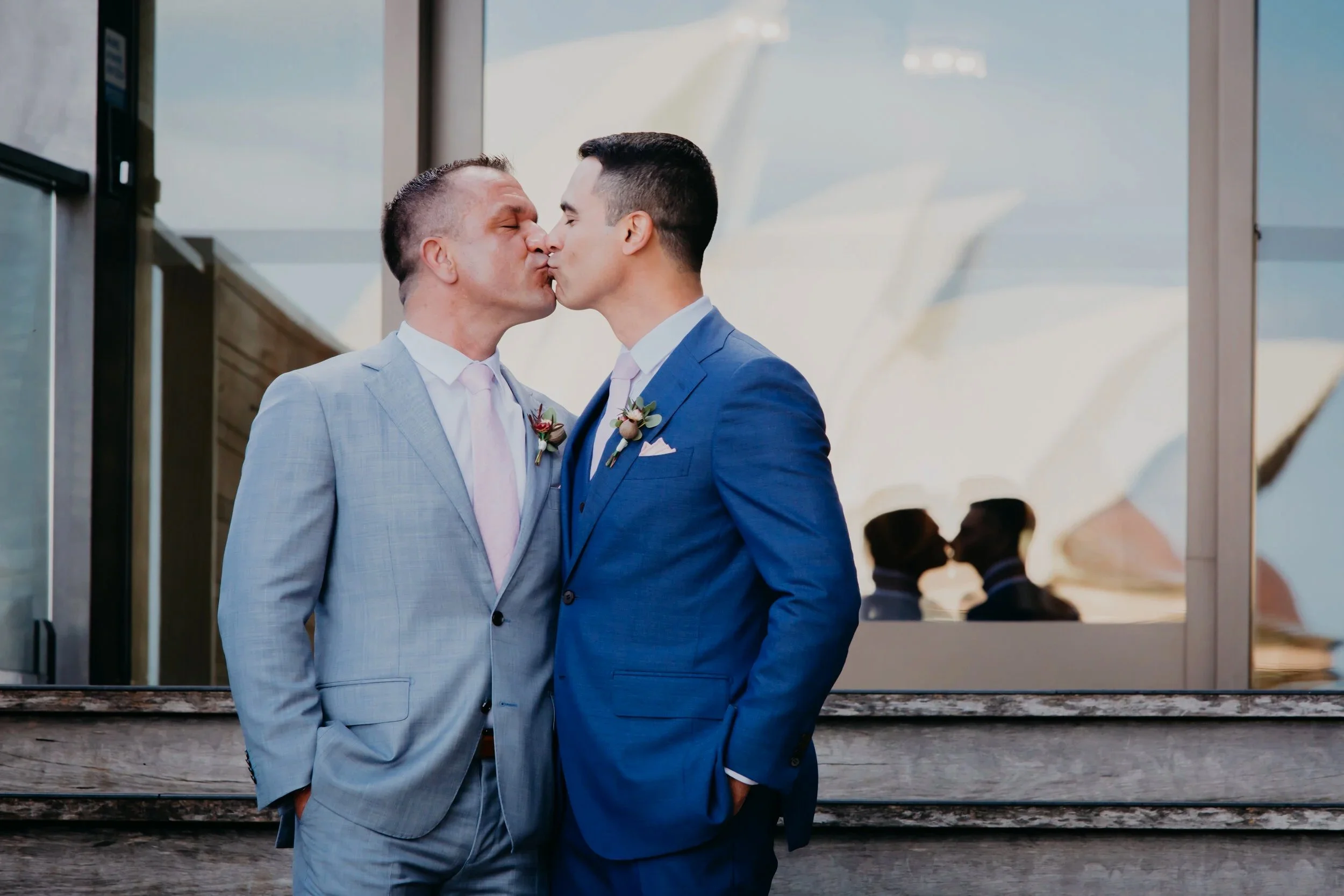 Two men in suits sharing a kiss at a wedding, with the Sydney Opera House in the background.