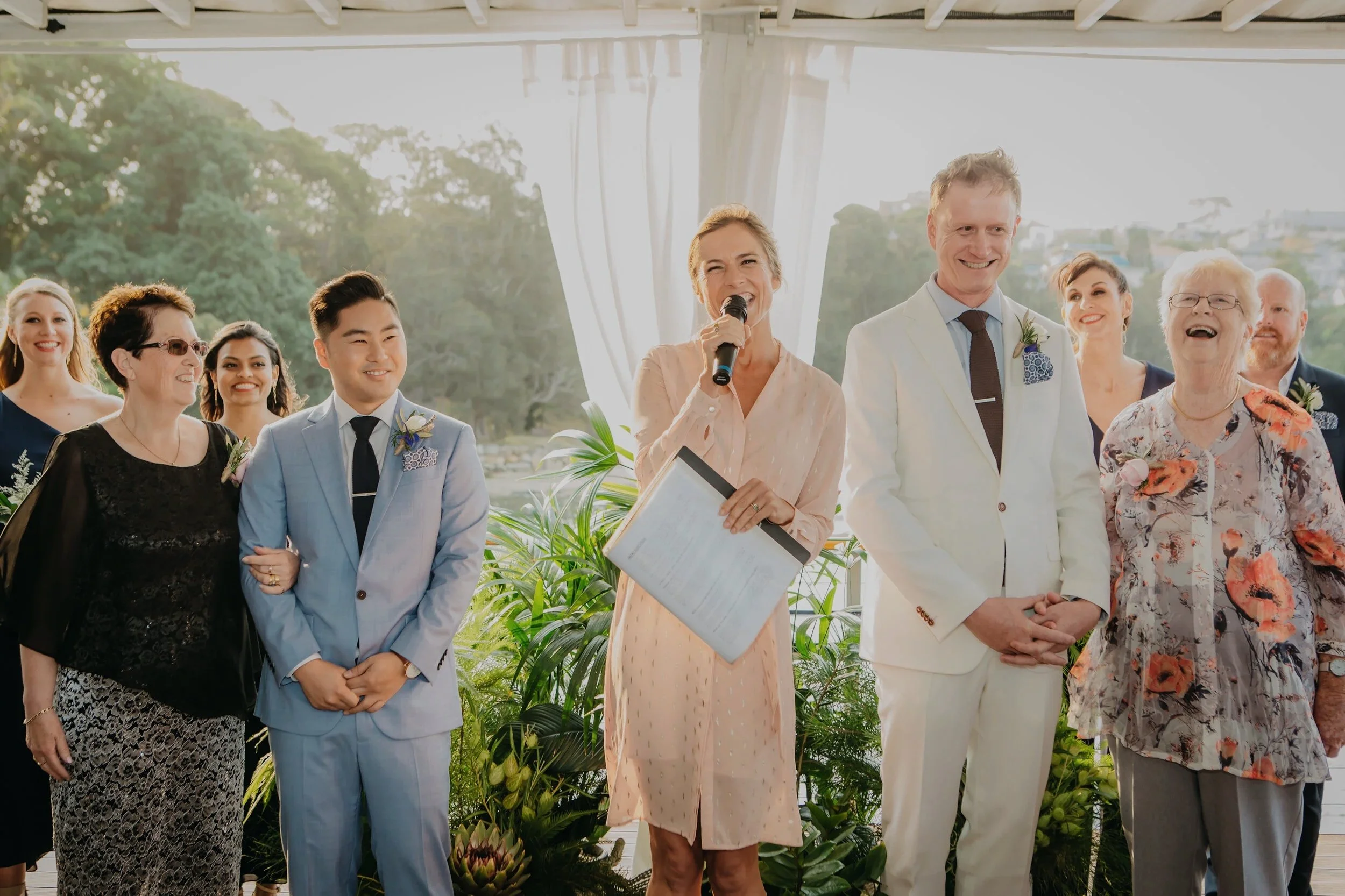 A group of people standing together outdoors, dressed in formal attire, at a wedding celebration. The woman in the center is speaking into a microphone, holding a script, with the bride and groom beside her, all smiling.