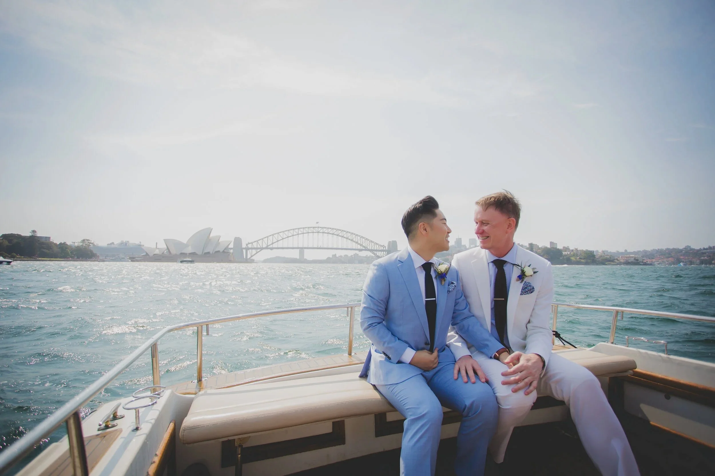 Two men in suits sitting on a boat with Sydney Opera House and Harbour Bridge in the background, smiling at each other, during daytime.