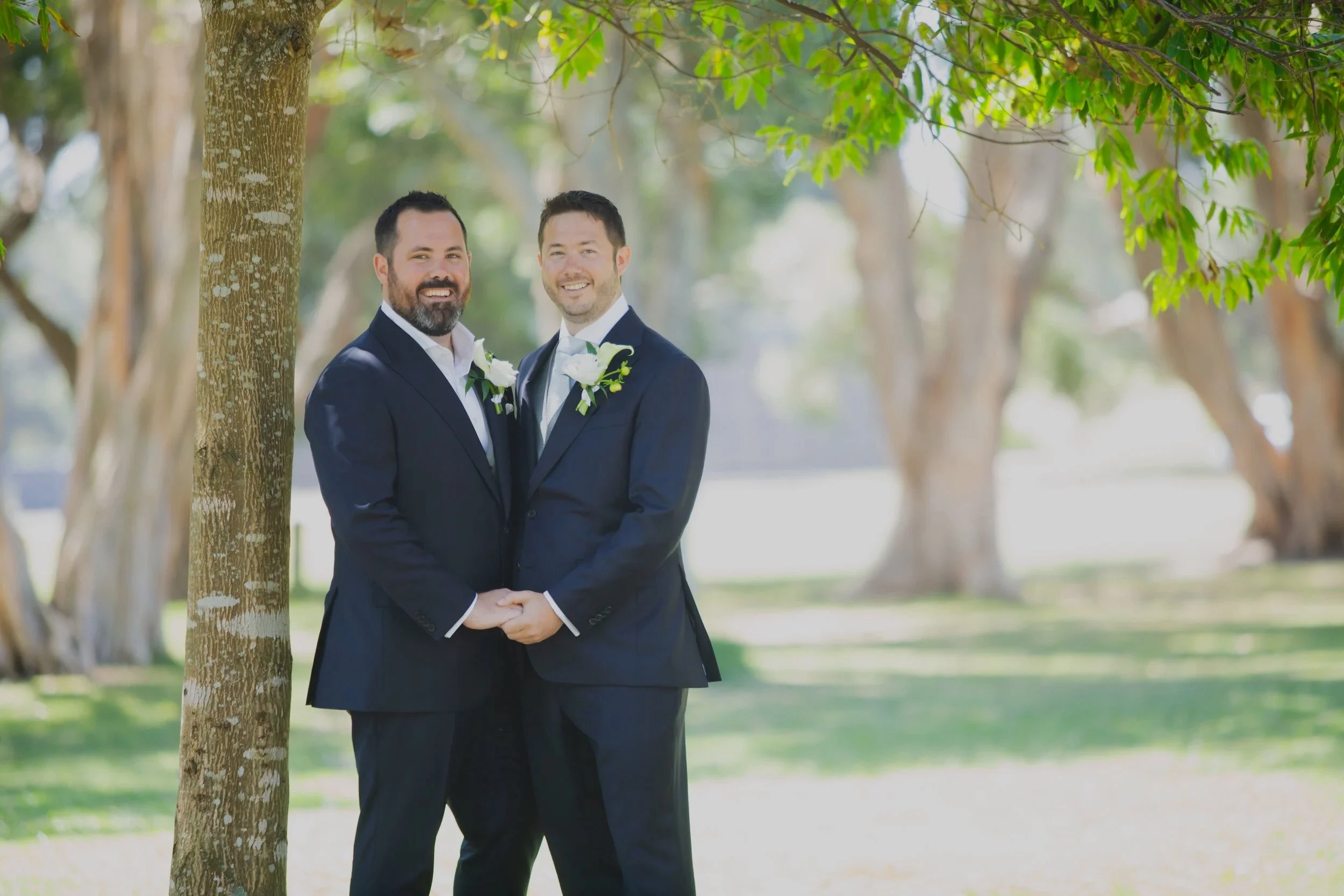 Two men in tuxedos holding hands and smiling outdoors on a sunny day, near a tree with green leaves, in a park setting with grass and additional trees in the background.