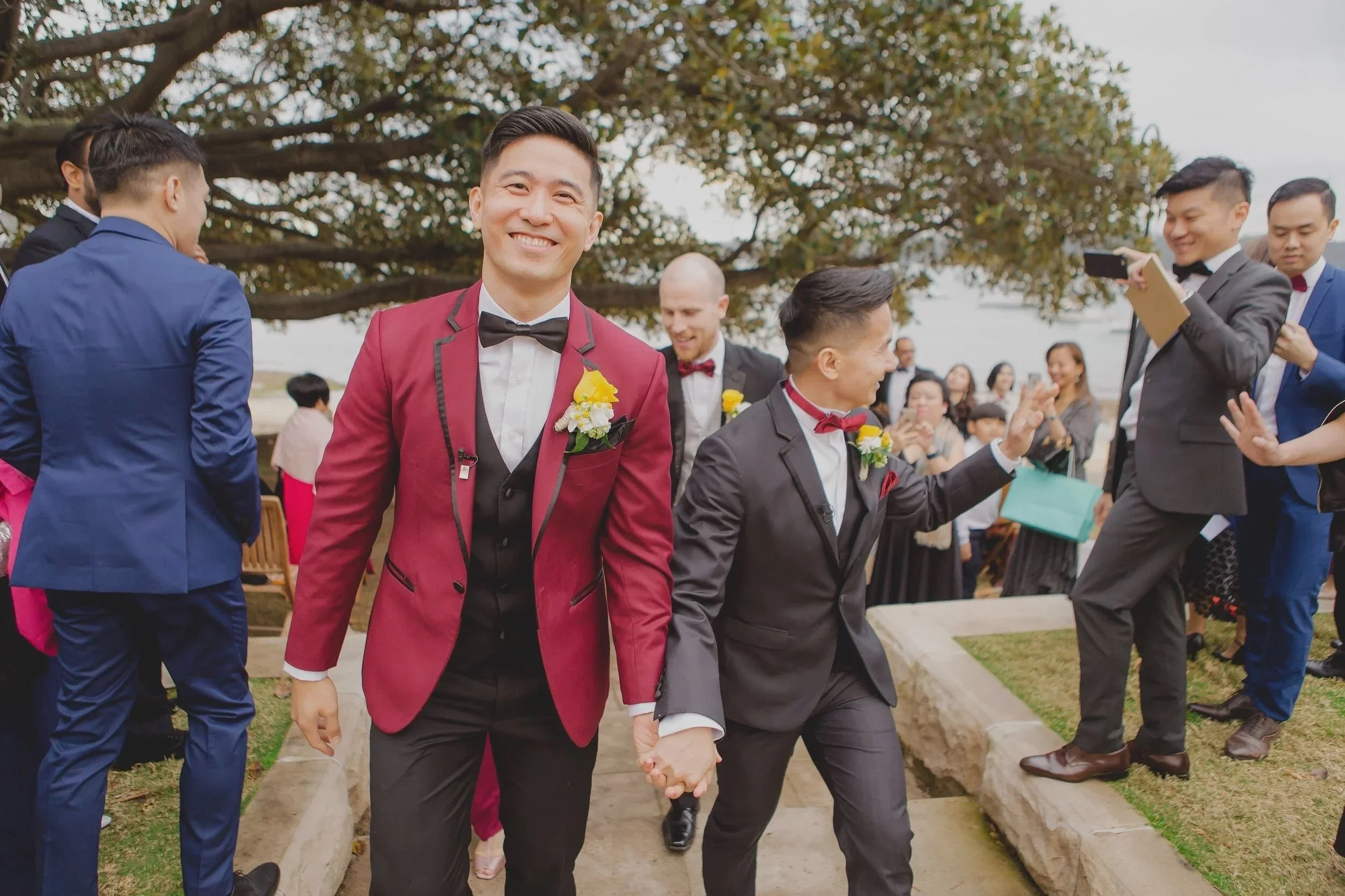 Gay couple in tuxedos holding hands and smiling at outdoor wedding ceremony with friends in the background.