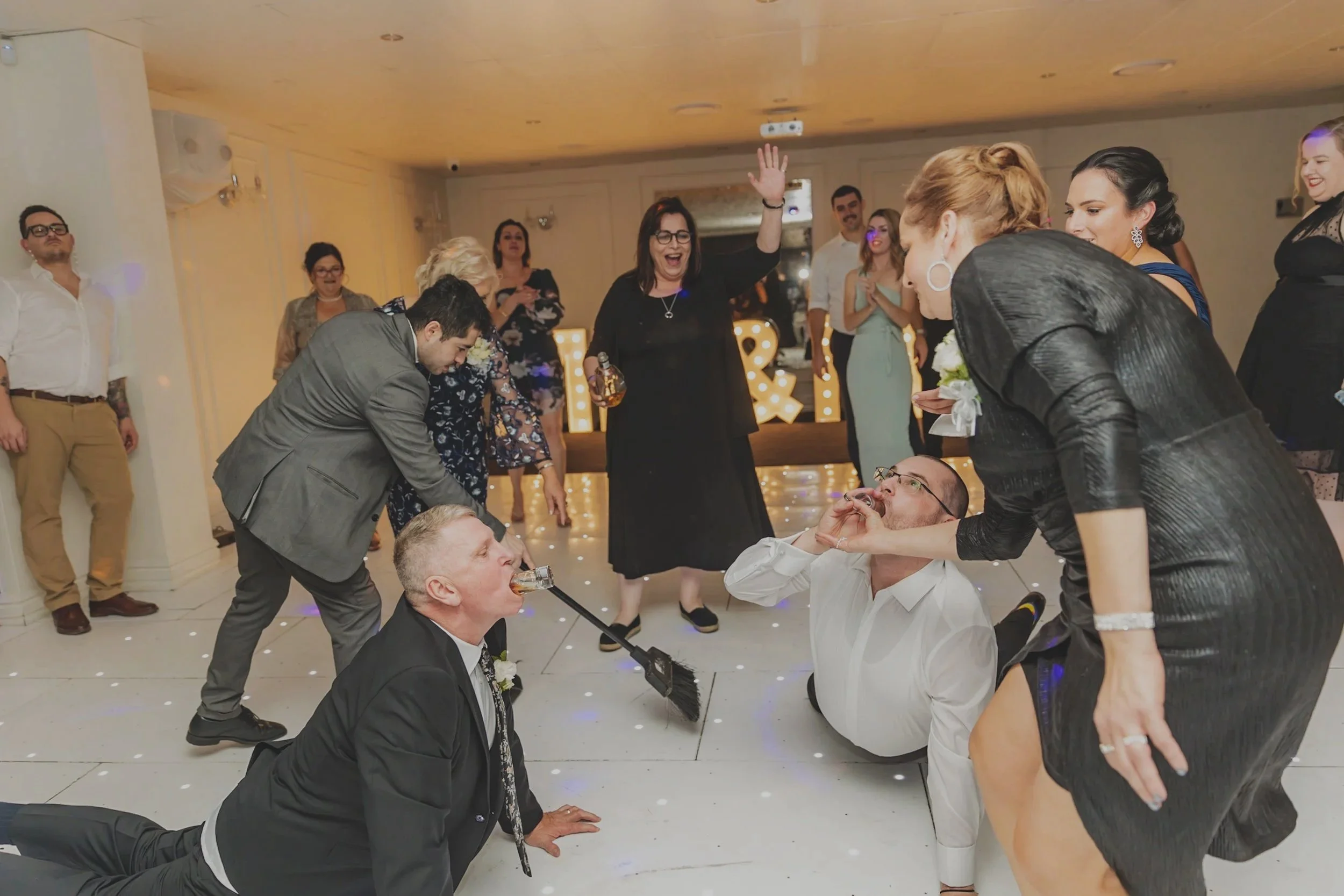 People dancing and having fun at a party, with some kneeling on the floor drinking shots, while others stand and watch, in a decorated venue with large illuminated letters in the background.