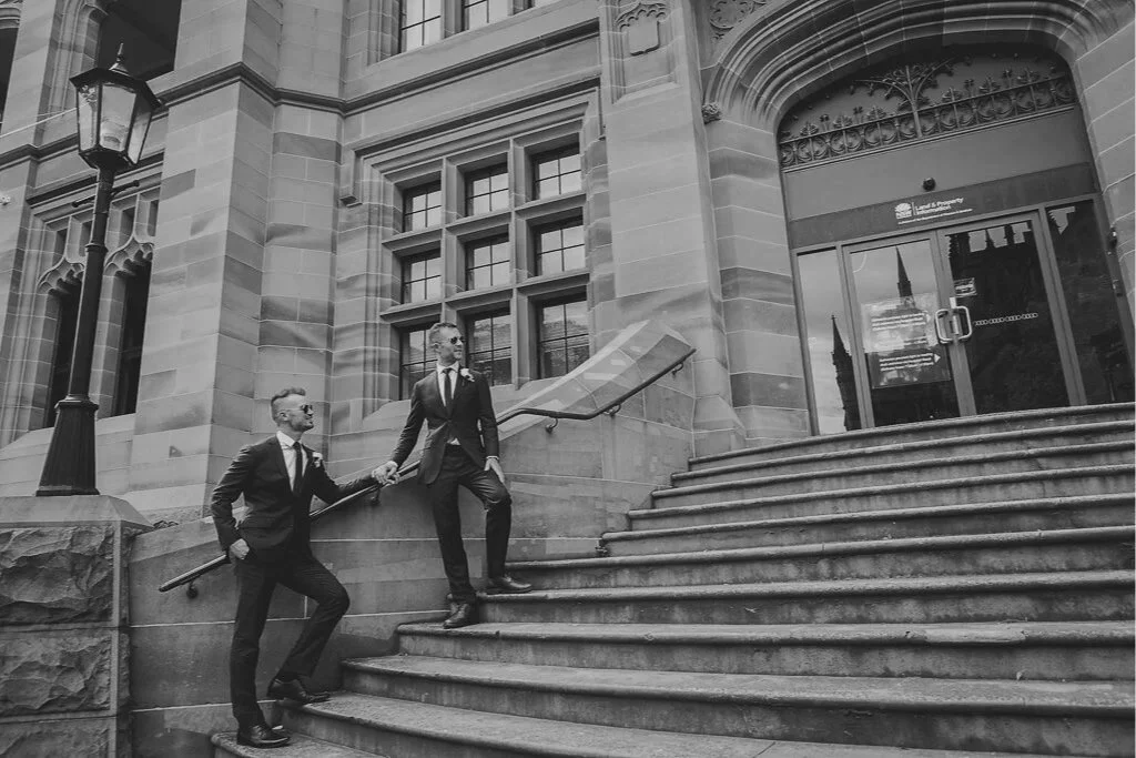Two men in suits on the steps outside a historic building with sandstone walls and large windows, holding hands.
