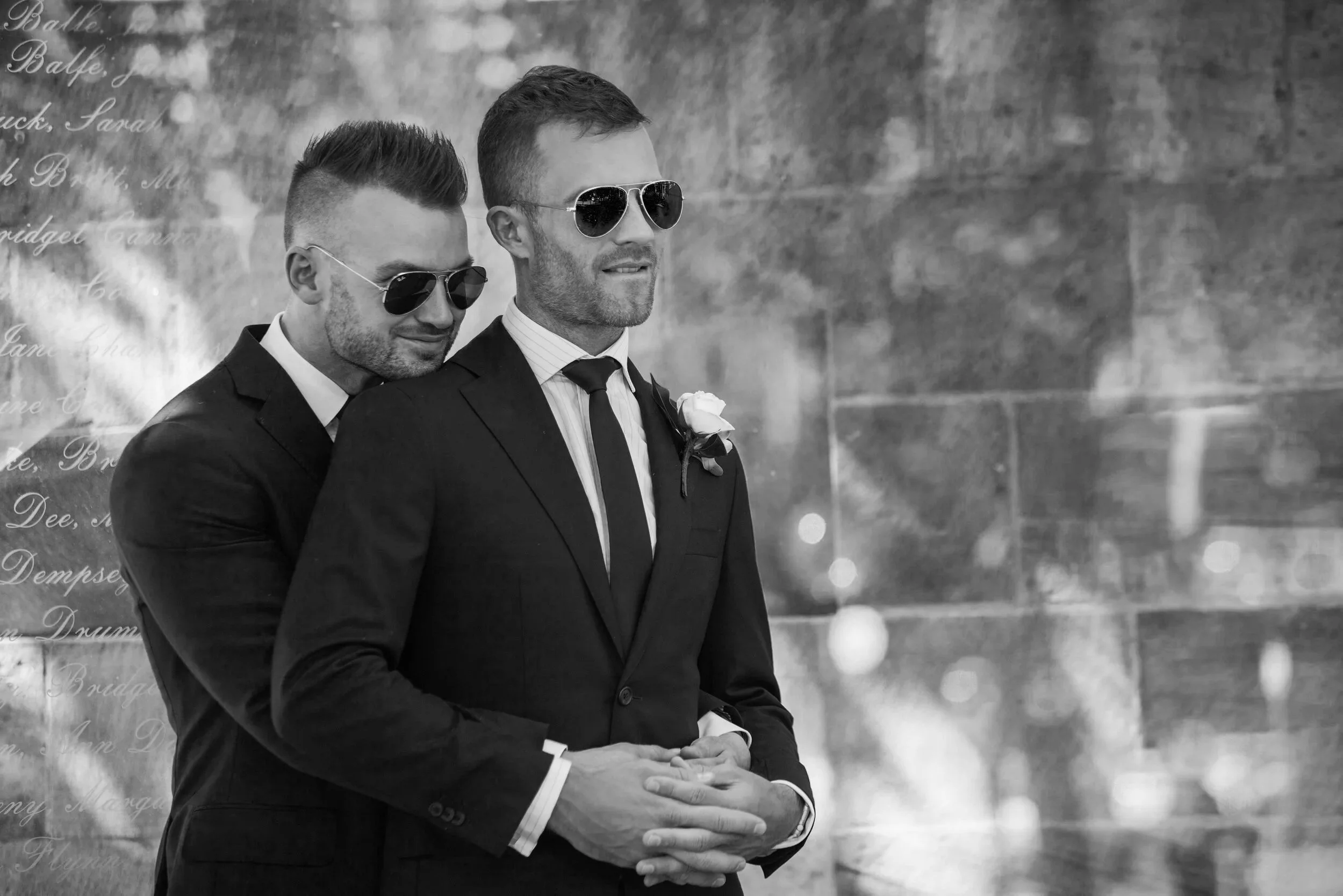 Two men dressed in suits, wearing sunglasses, one with a boutonniere, standing closely together outdoors during a wedding ceremony.