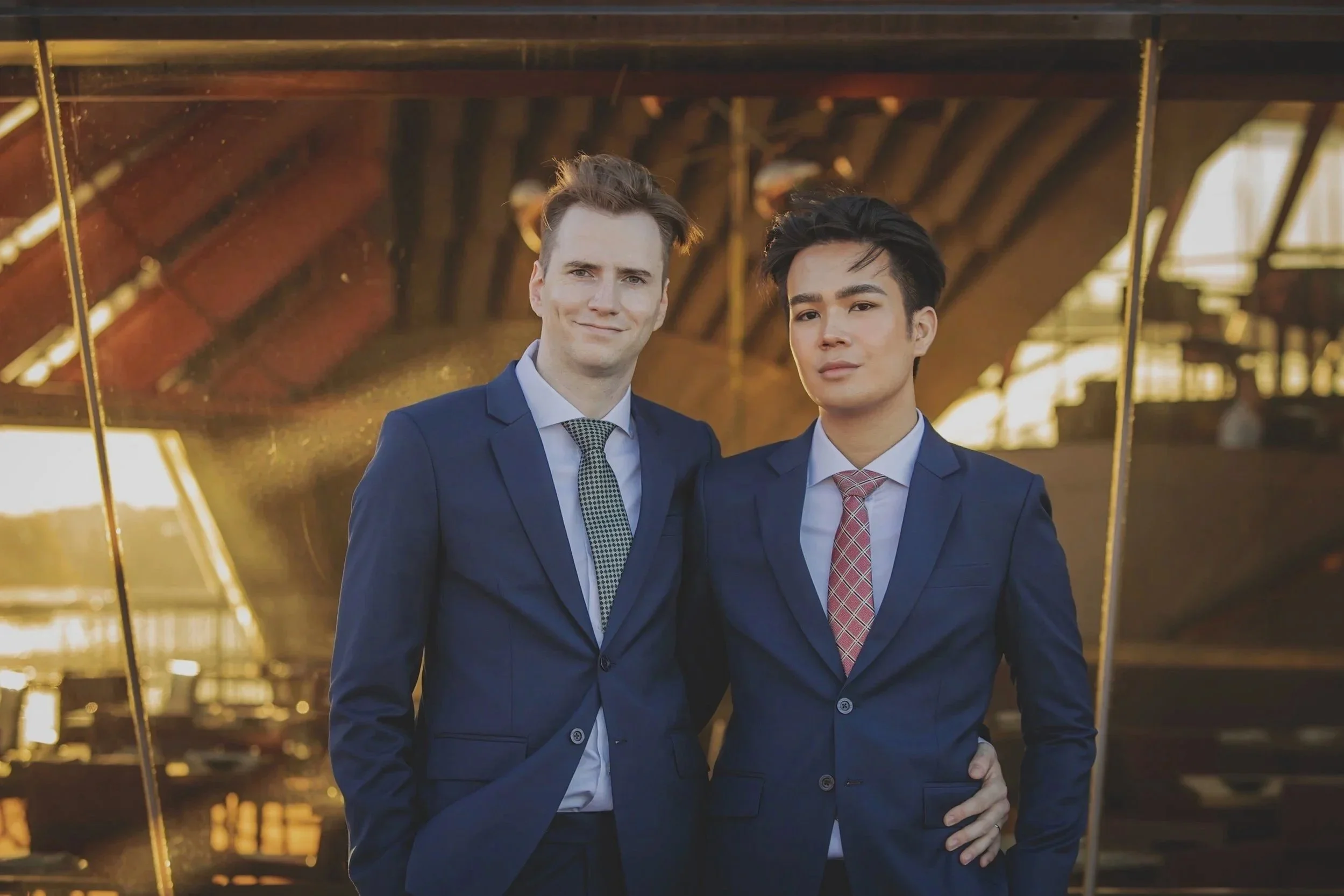 Two young men dressed in navy suits and ties standing side by side outdoors, smiling for a photo in front of metallic, reflective surfaces.