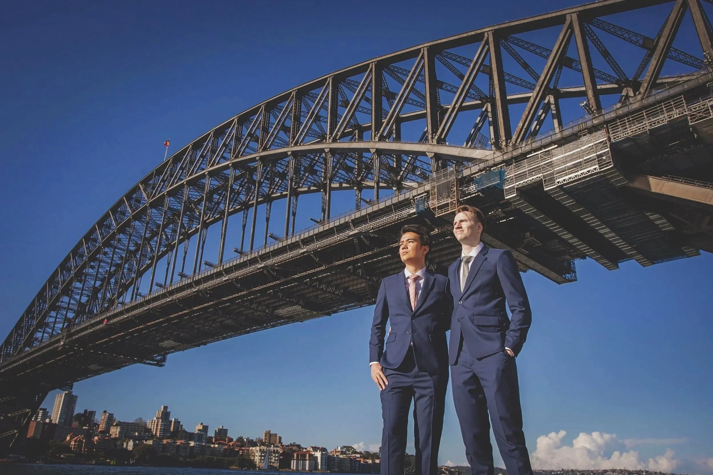 Two businessmen in suits standing under Sydney Harbour Bridge with city buildings in the background on a clear day.