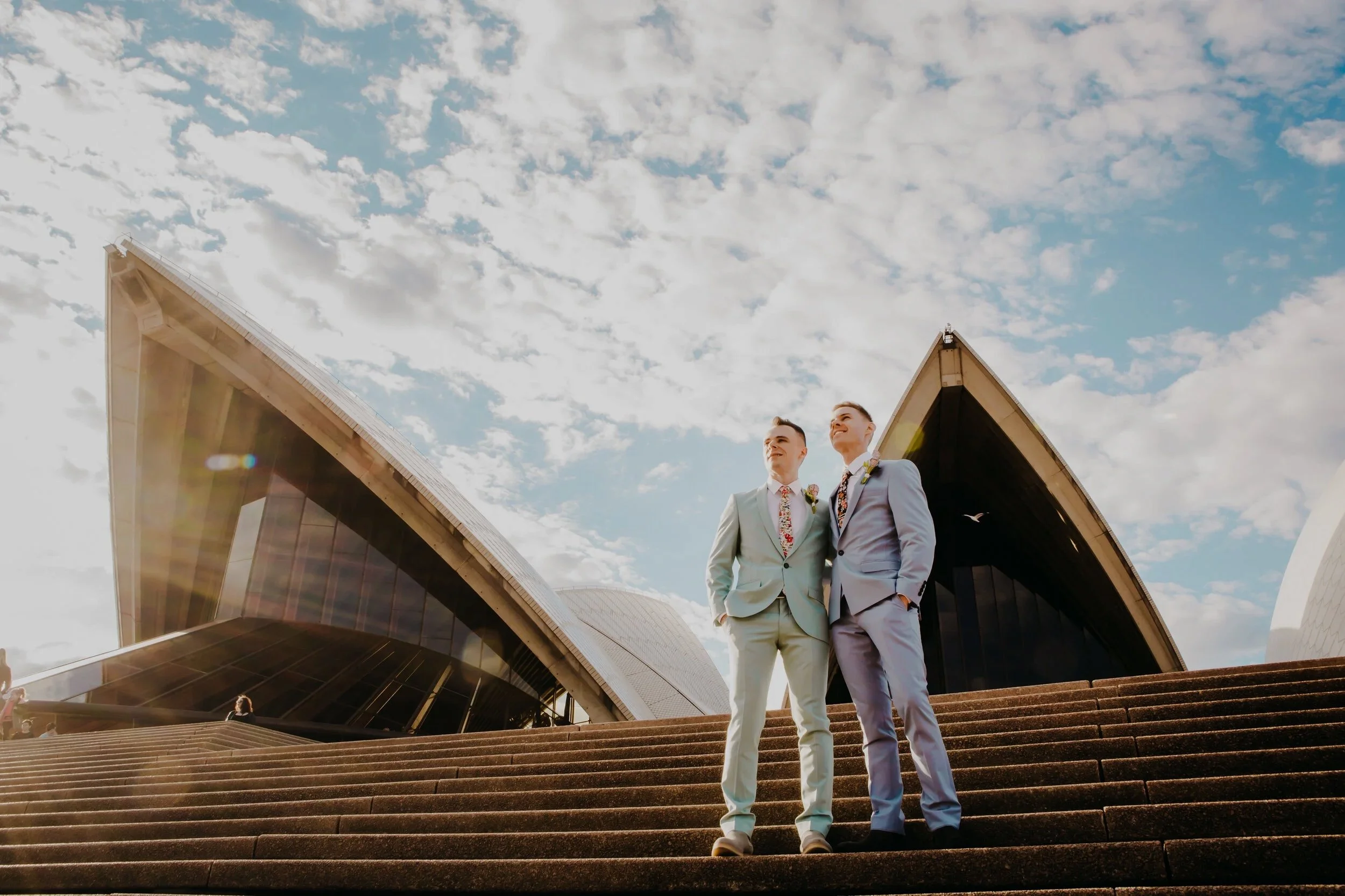 Two men in suits standing on steps outside Sydney Opera House, looking into the distance, with a partly cloudy sky overhead.