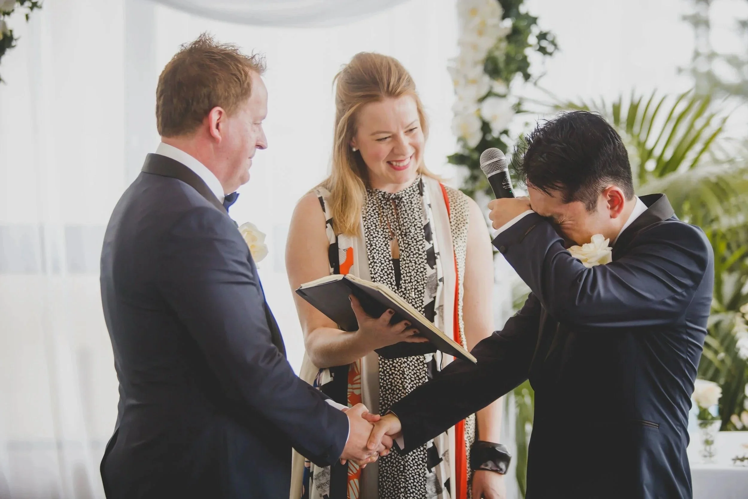 An emotional moment during a wedding ceremony where a groom is crying and holding hands with the bride, while a woman officiates, holding a book and a microphone, and there's a floral background.